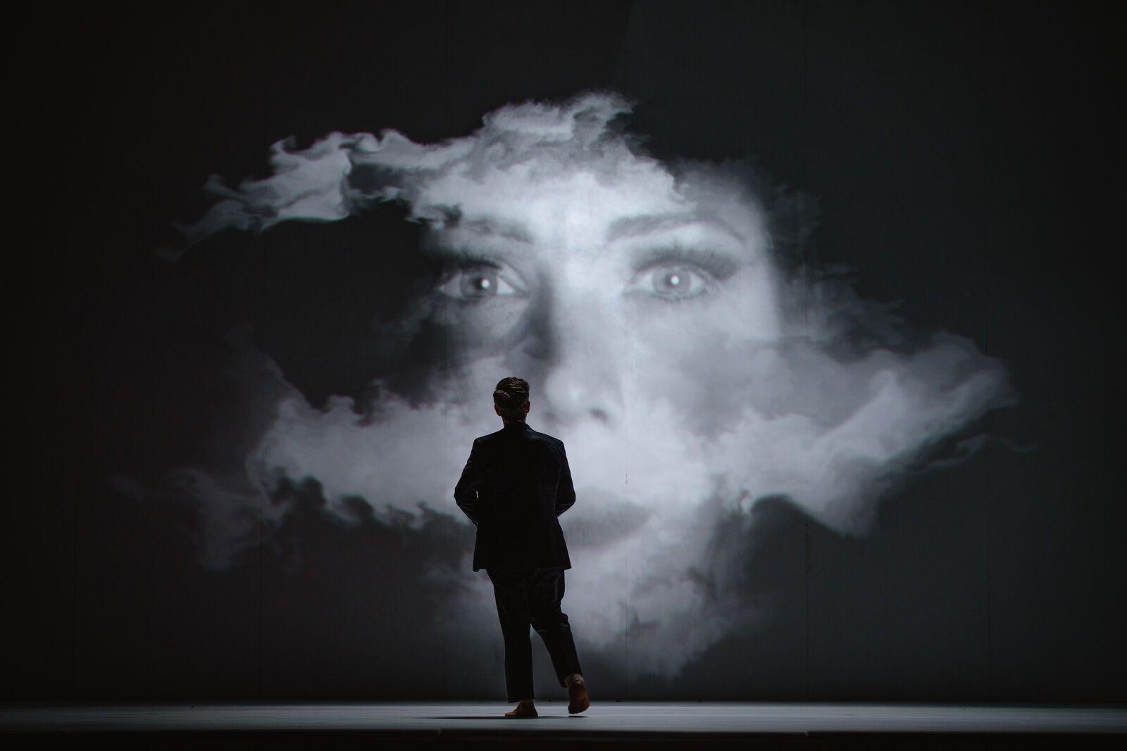 There is a silhouette of a man looking at a picture projected onto a wall of a woman looking through clouds. 