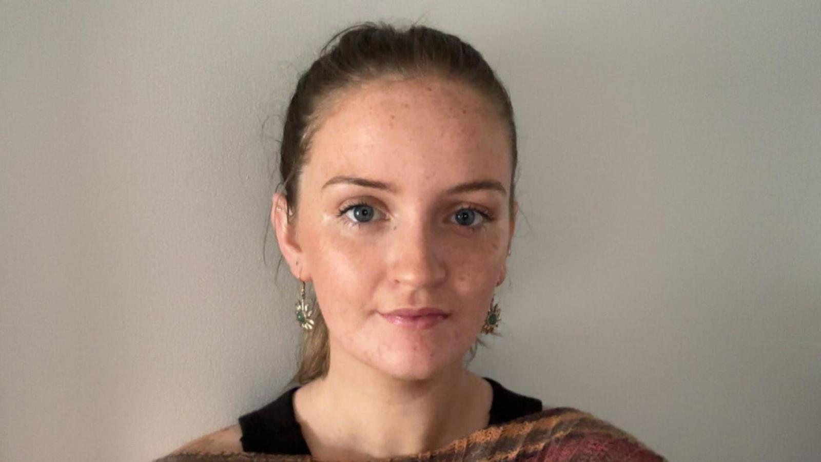 A blond young women with dangly gold earrings and blue eyes against a white wall with her hair tied up.