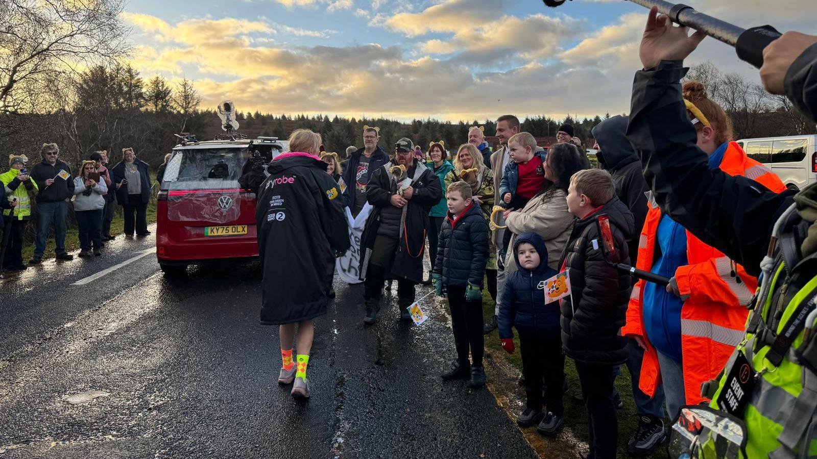 A woman facing away from the camera wears a big black jacket, colourful socks and trainers. Adults and kids stand around on either side of the road smiling at her. A number of them wear headbands will yellow ears on them. 