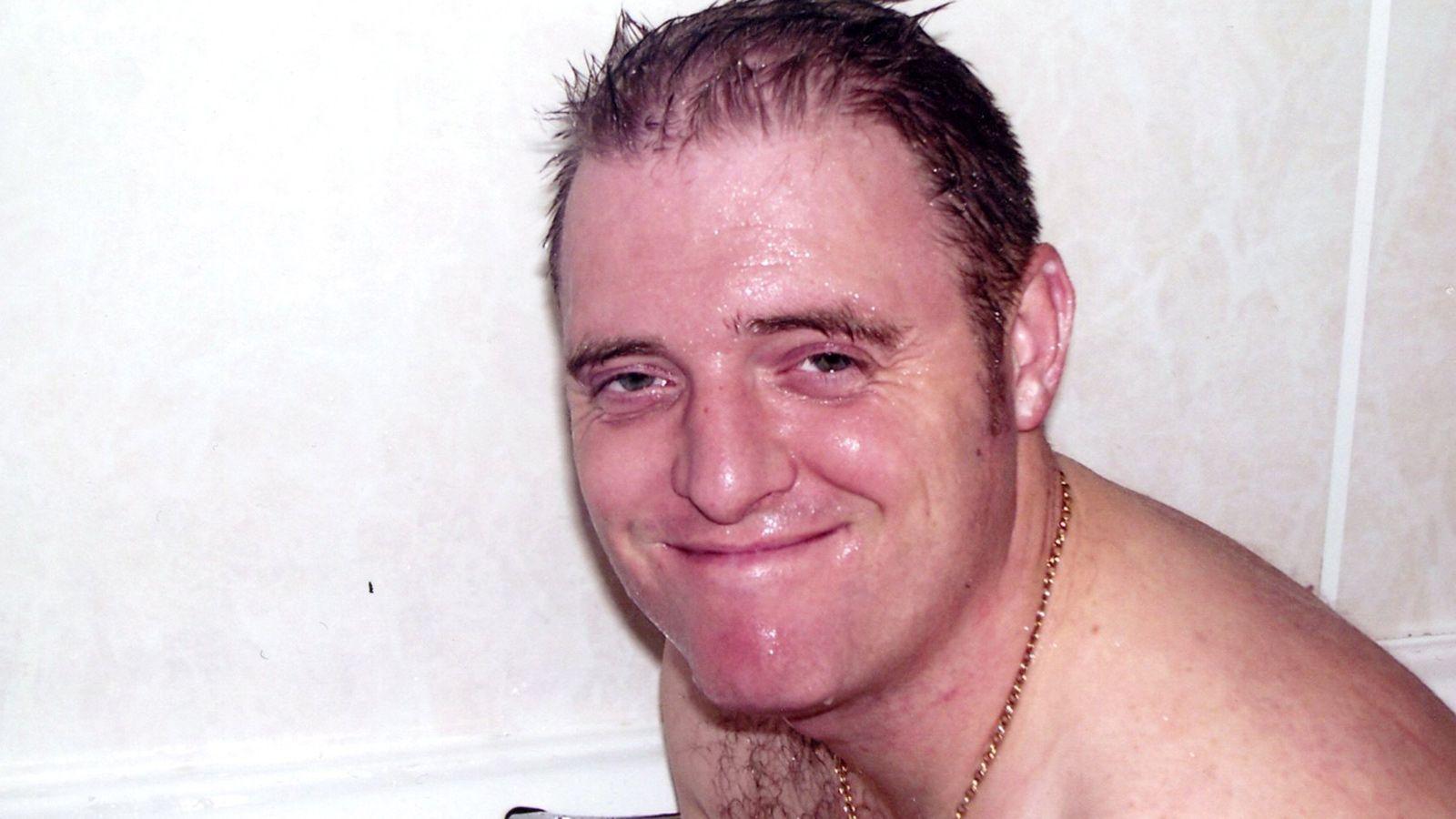 A wet man with short, dark, receding hair sits in a bath grinning at the camera. He is wearing a gold necklace.