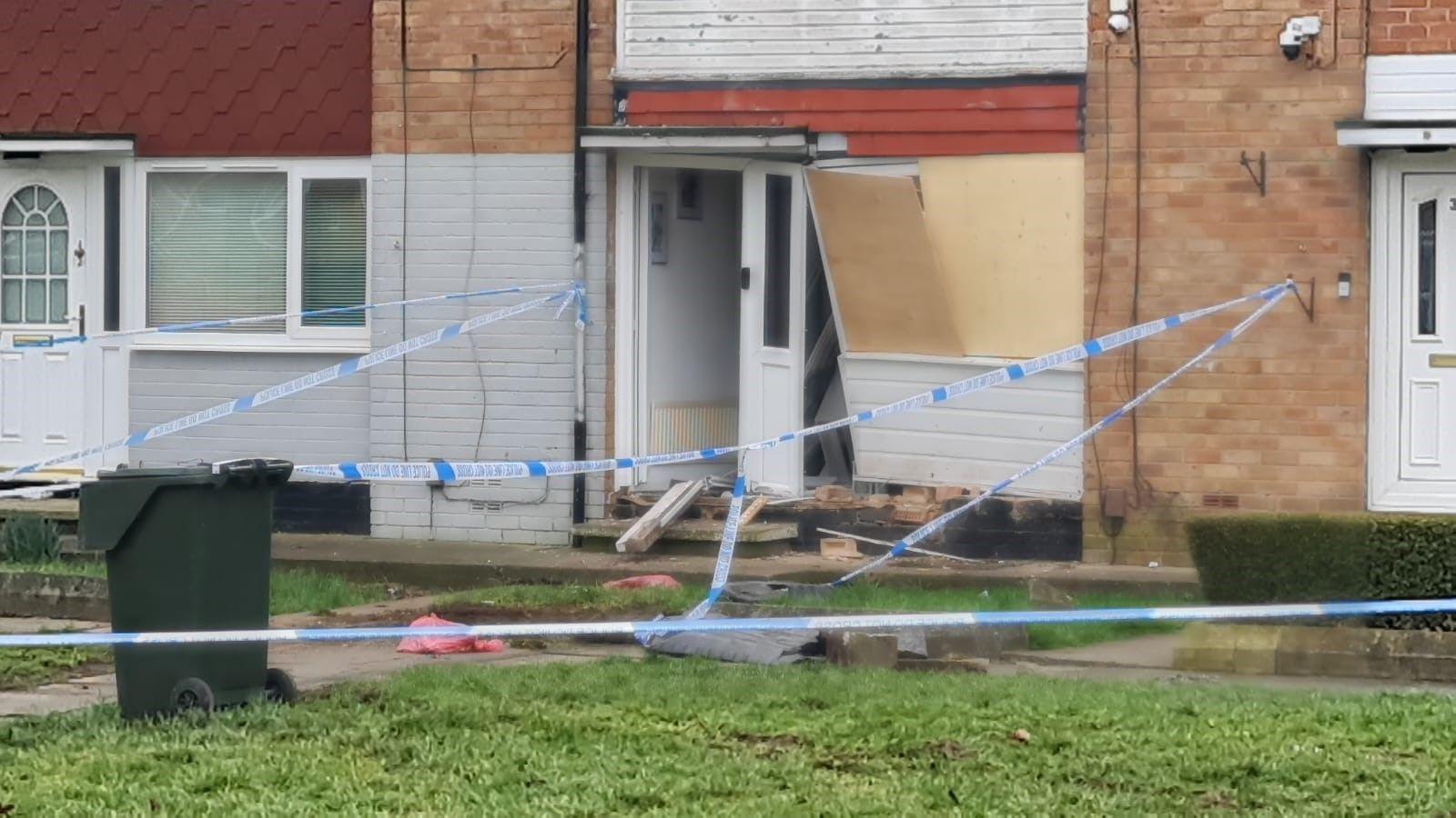 A terraced house cordoned by police tape with the front door and window severely damaged.