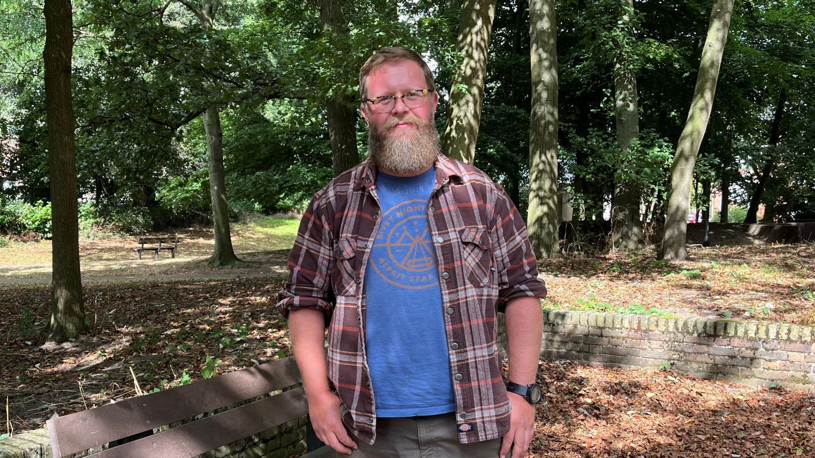 Man with a long beard wearing glasses and a check shirt and standing in a wooded area. 