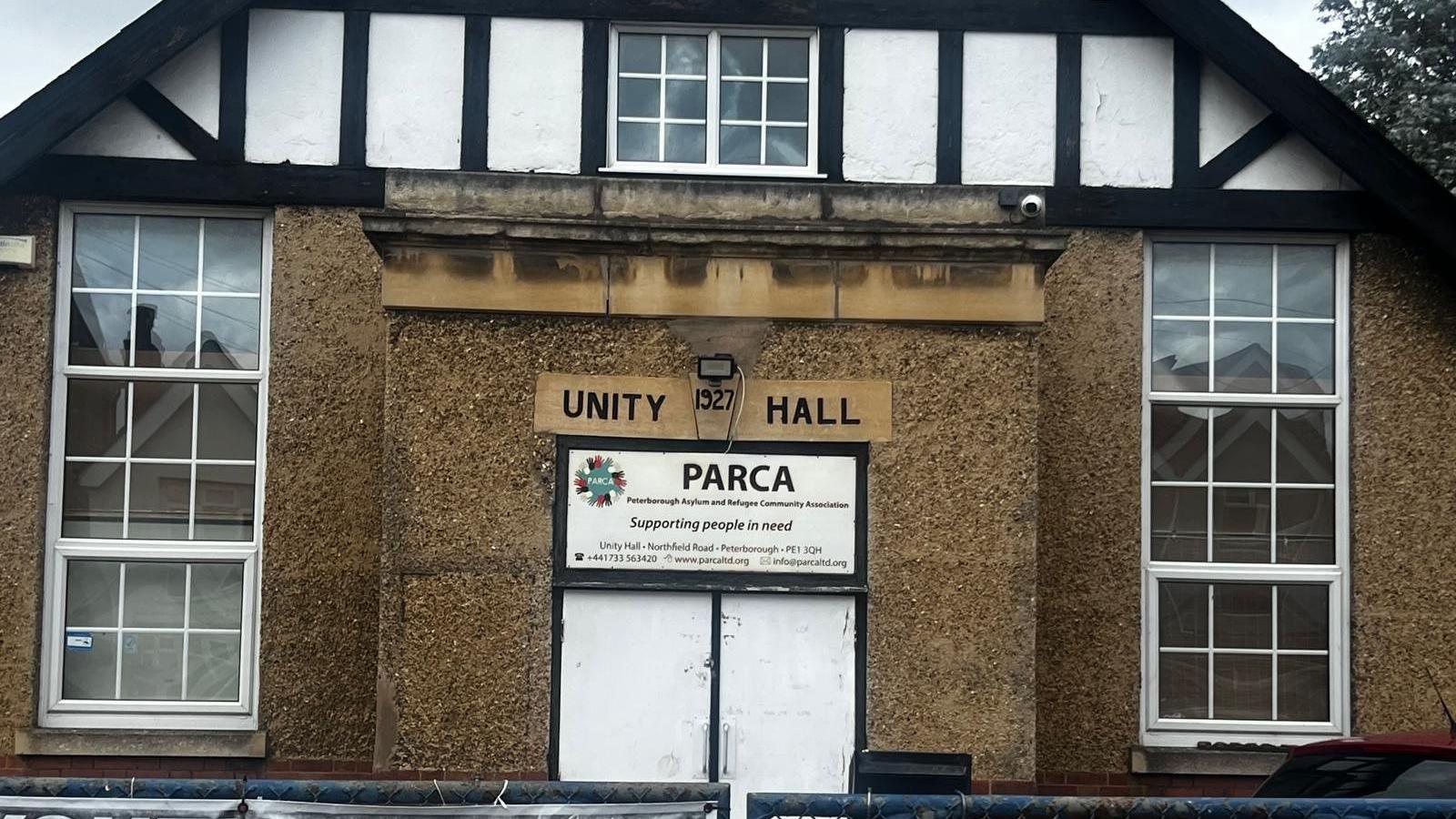 The exterior of a building, showing three windows with white window panes. The front is a mixture of brown stucco and at the top is white stucco with black beams. There is a sign that reads "Unity Hall 1927" and beneath it is a sign that reads "PARCA: Supporting people in need".