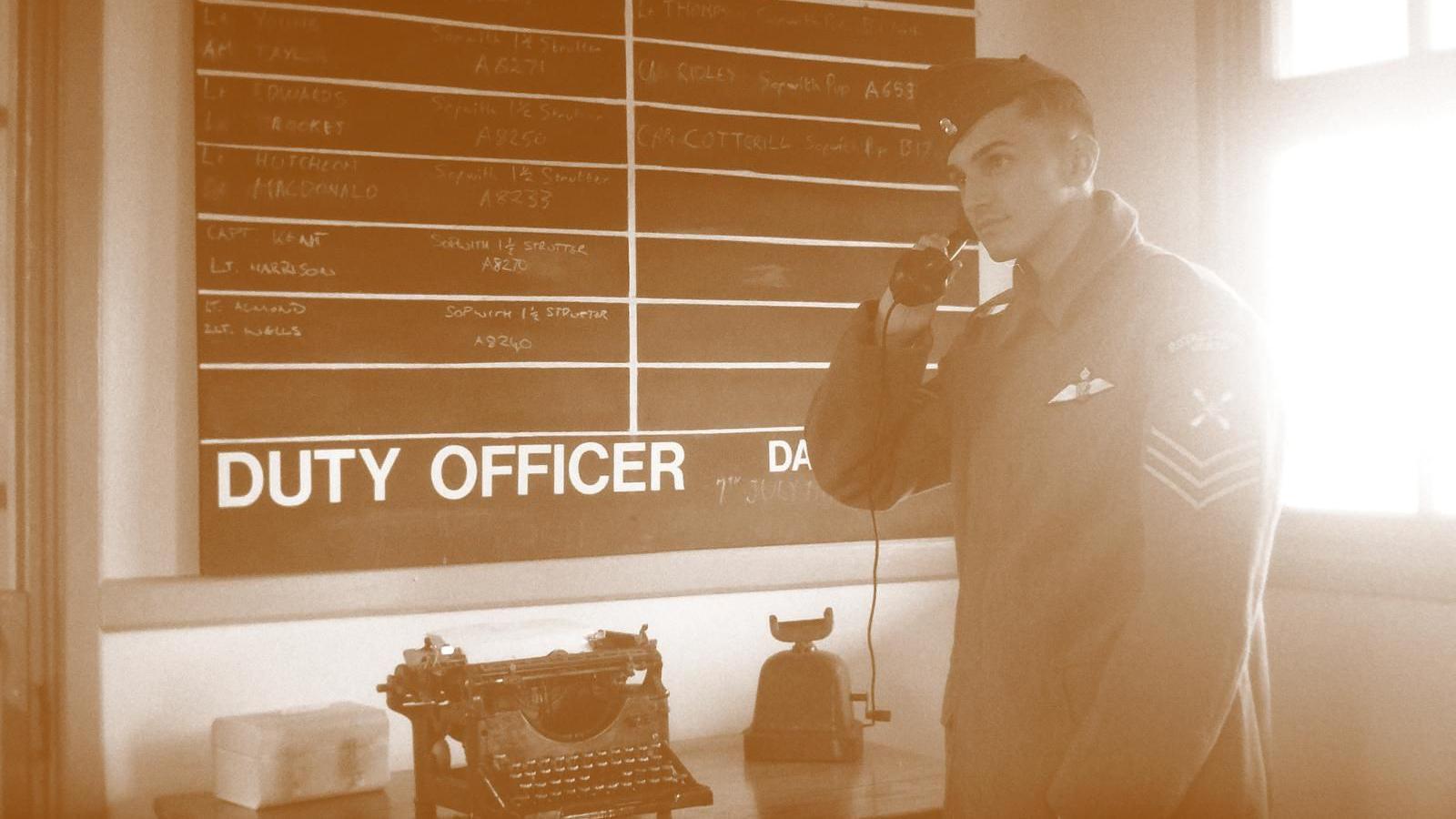 A sepia-coloured photo of a man in a soldier's uniform with three stripes on his arm, he is holding an old-fashioned phone to his ear and looking into the distance