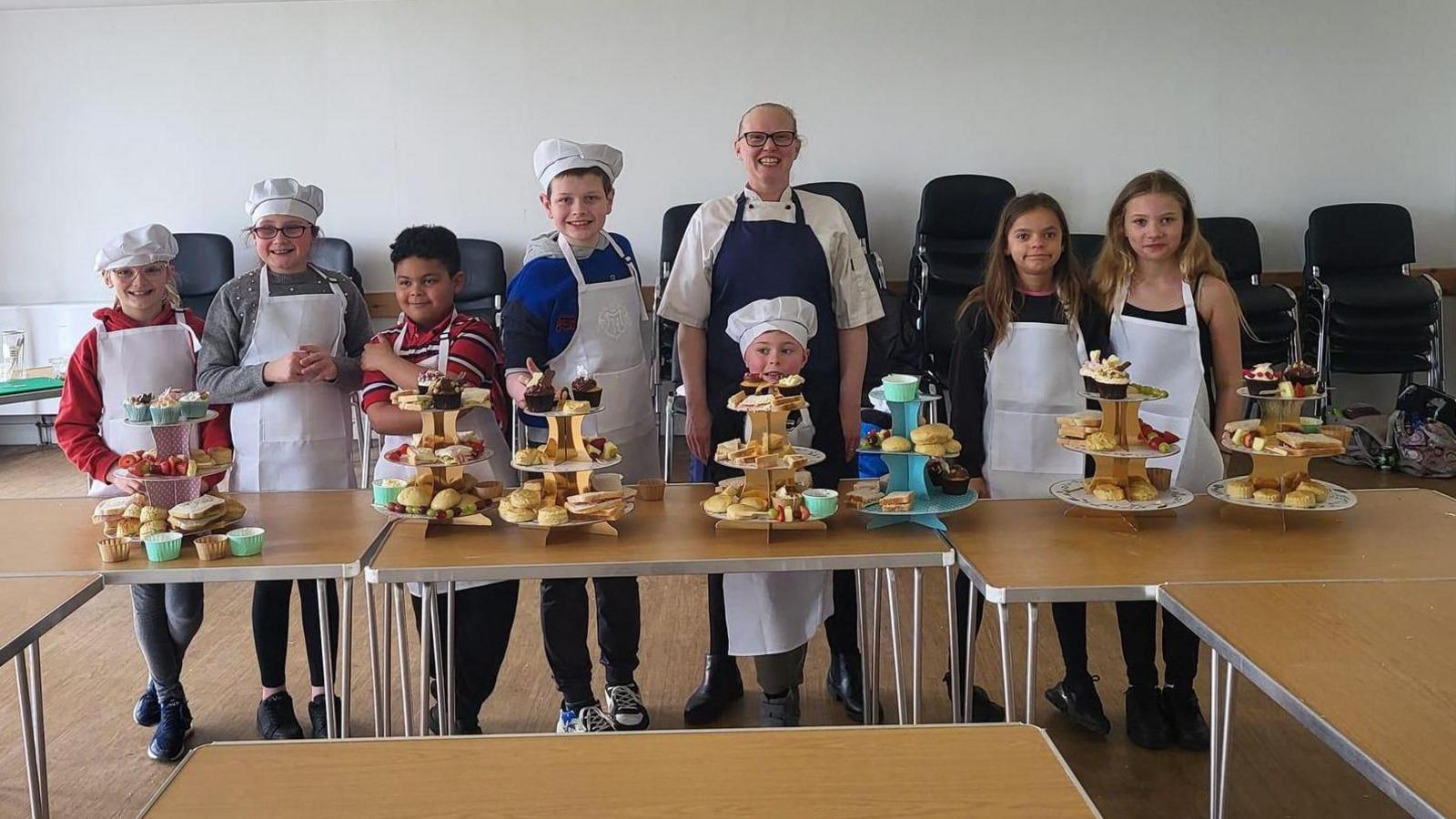 Seven children and one adult wearing white aprons and chefs hats standing in front of multi-tiered afternoon tea stands with cakes and sandwiches on. They're in what looks like some kind of community hall, with chairs stacked up behind them.