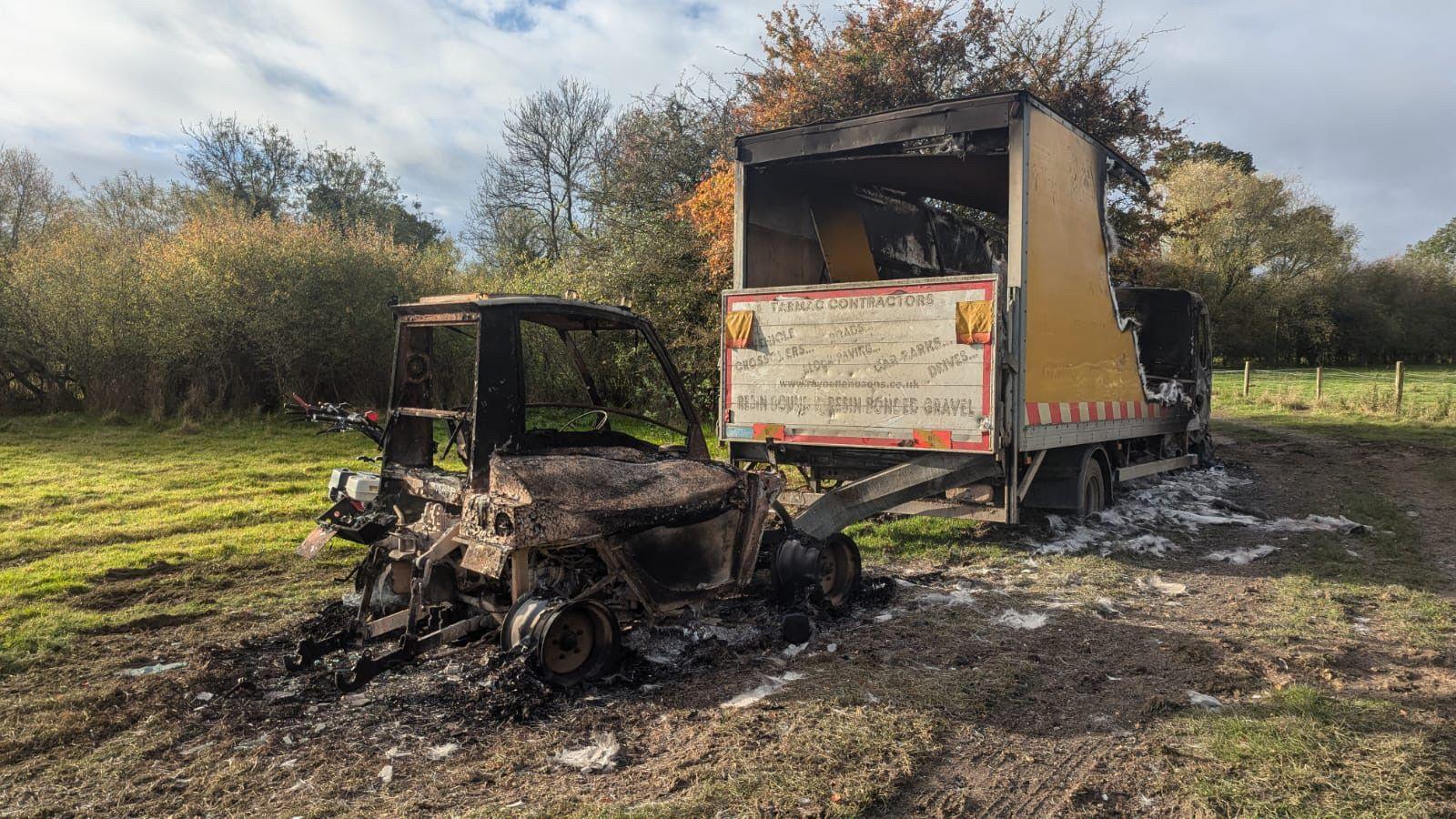 The burned shell of a lorry on a muddy surface with a field in the background and a smaller vehicle behind it, also burned