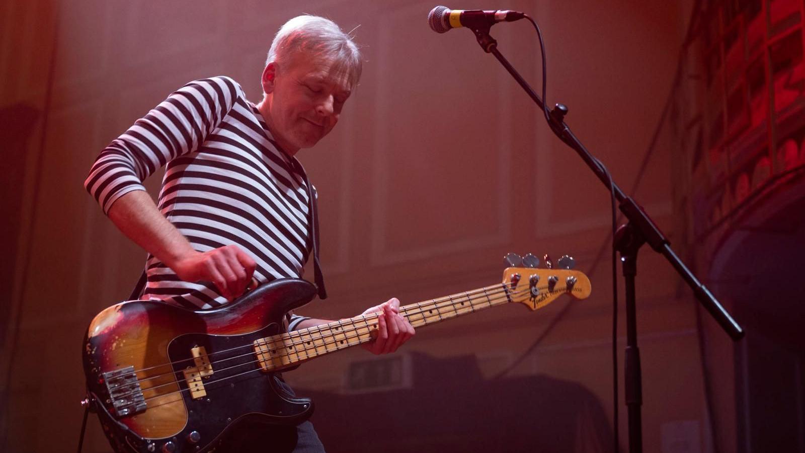 Rob Tickell is performing on stage with an electric guitar. He is wearing a striped shirt and smiling.