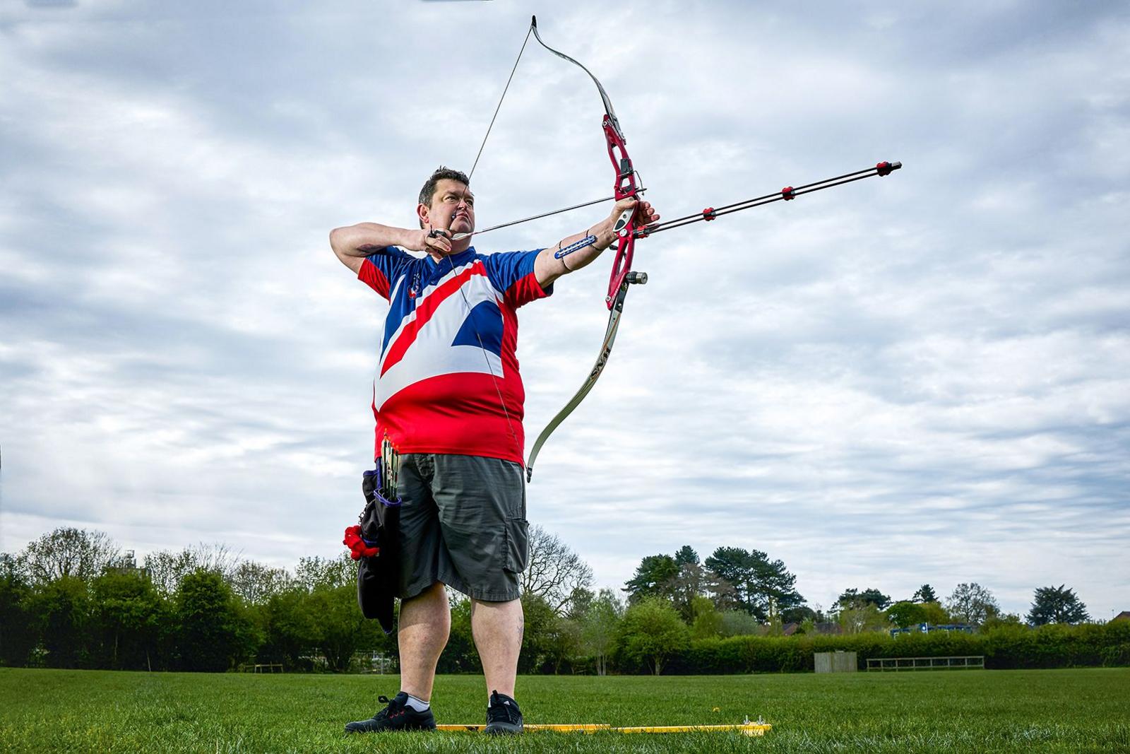 A man with short brown hair wearing a blue, white and red sports t shirt and khaki shorts. He is holding a bow and arrow, as though he is about to shoot and is stood in a large graddy field with trees in the background.