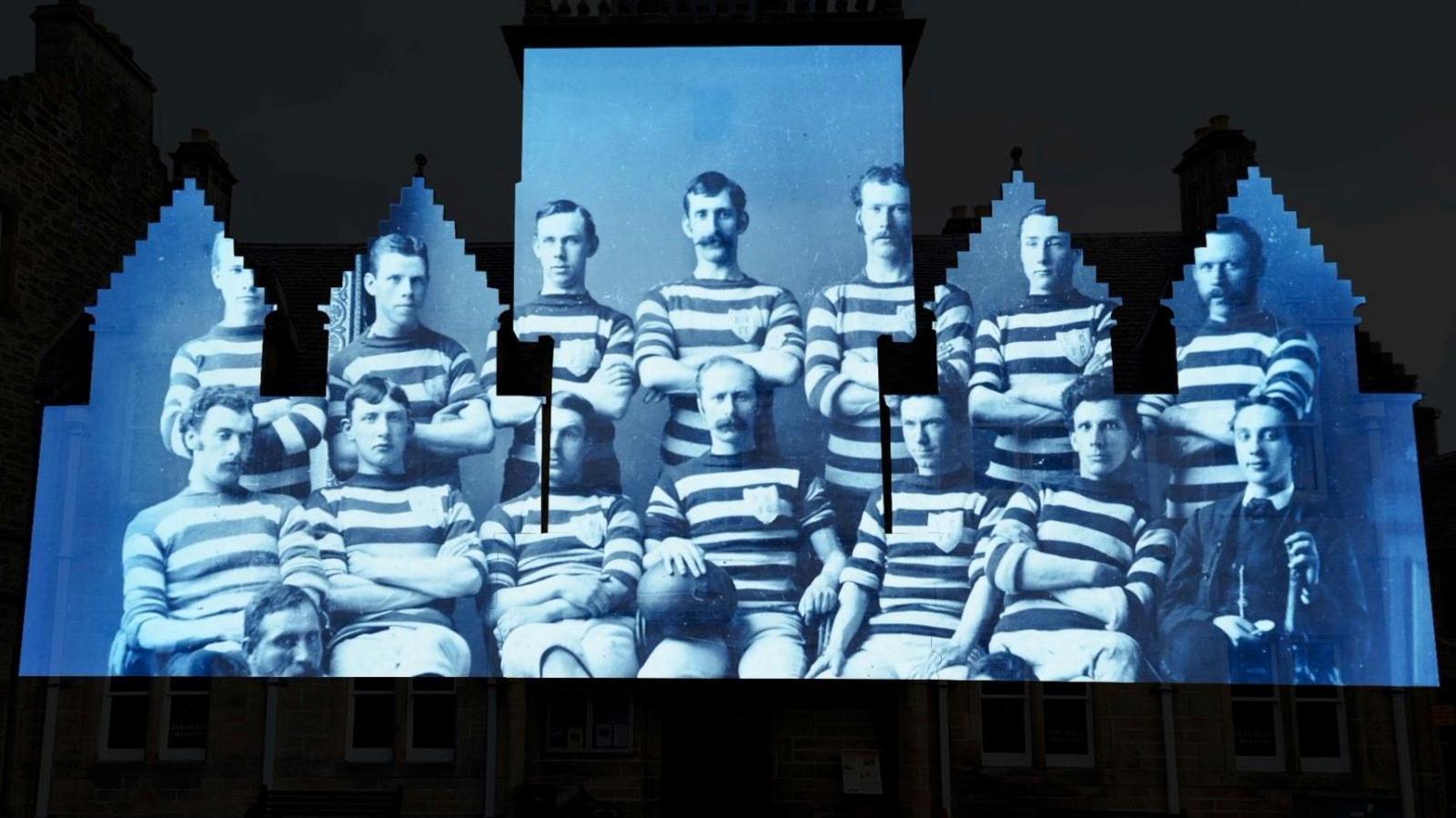 The projection is blue in colour and shows an archive image of a football team, the players wearing hooped shirts. Some of the men are sporting large moustaches.