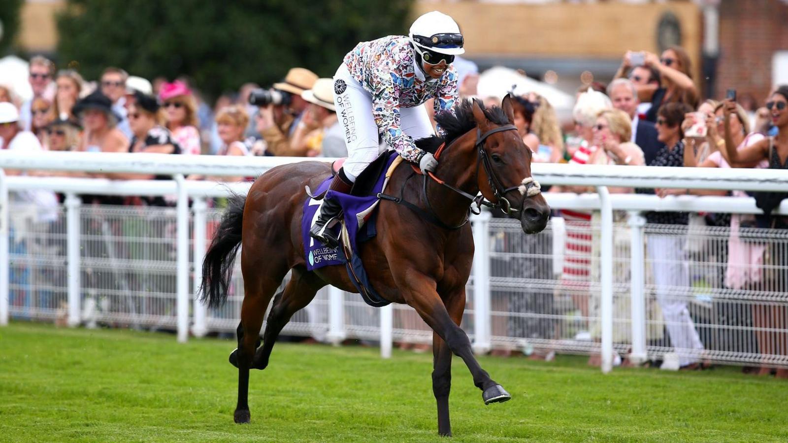 A woman wearing a hijab and a helmet and a colourful horse-riding top. She is riding a horse. There is a crowd in the background. 