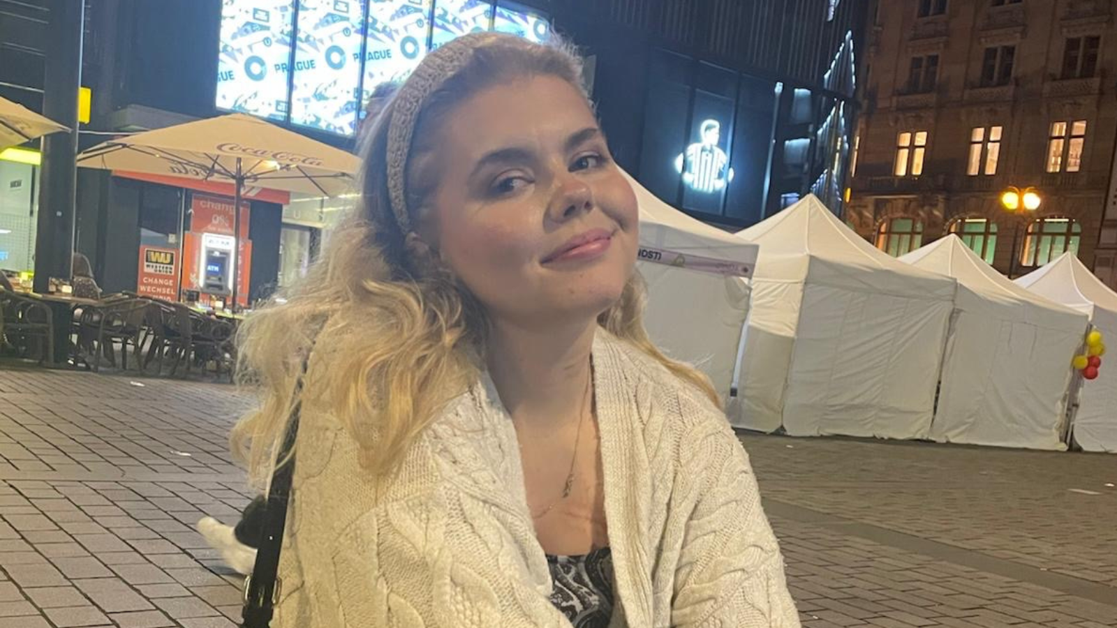 Anya Roberts smiles at the camera, in a market square. She has blonde wavy hair and a white headband, and wears a white cardigan and a patterned top.