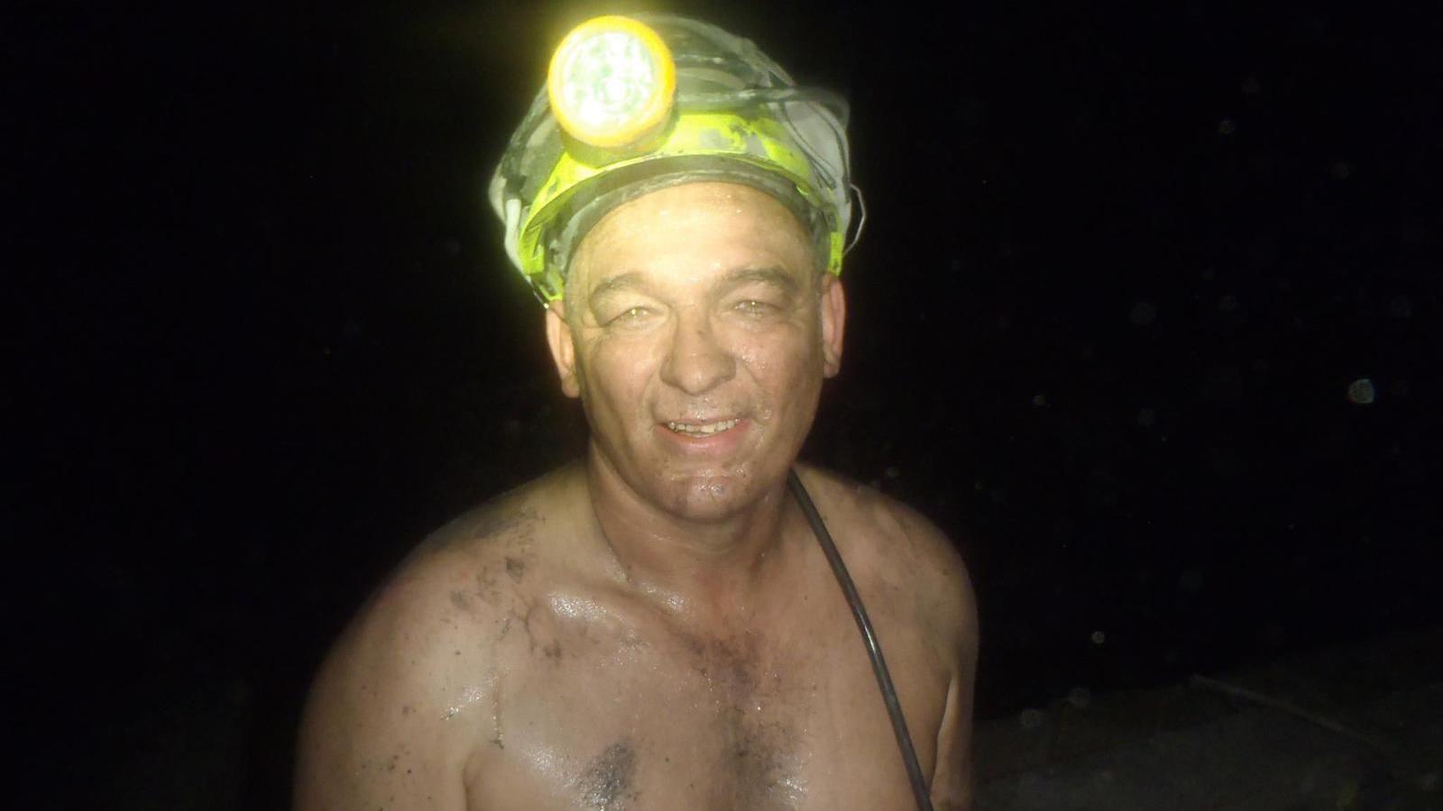 Garry Owen, bare-chested and smiling, down a coal pit with a helmet and torchlight on his head. 