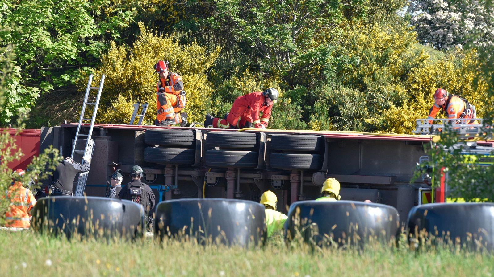 the underside of a lorry can be seen after it overturned. there are a number of rescue workers on top of the lorry and standing to its side. The area is surrounded in green bushes, trees and long grass.