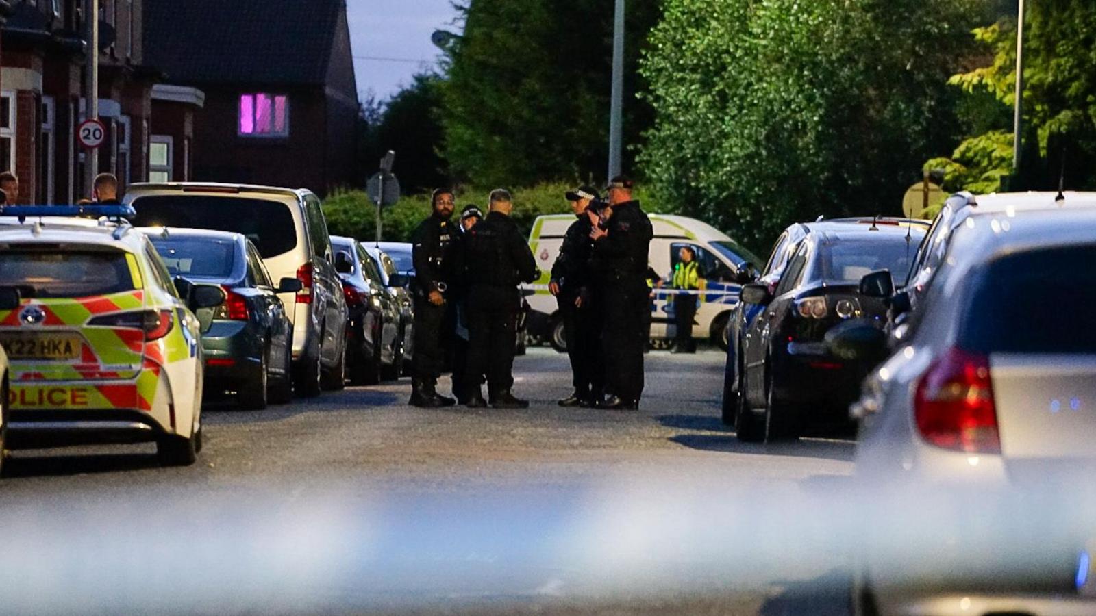 A number of police officers in uniform stand between parked cars on a terraced street, with police tape across the road in the background and a police van