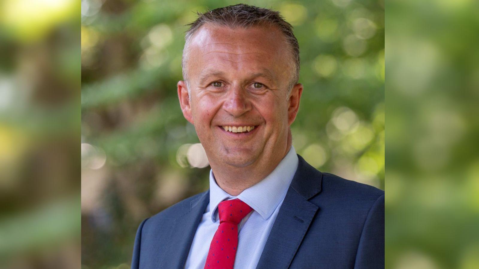 A professional headshot of Will Penny wearing a light blue collared shirt, a red tie and a navy blazer. He has dark grey hair and is standing outdoors in front of trees, which are blurred in the background.