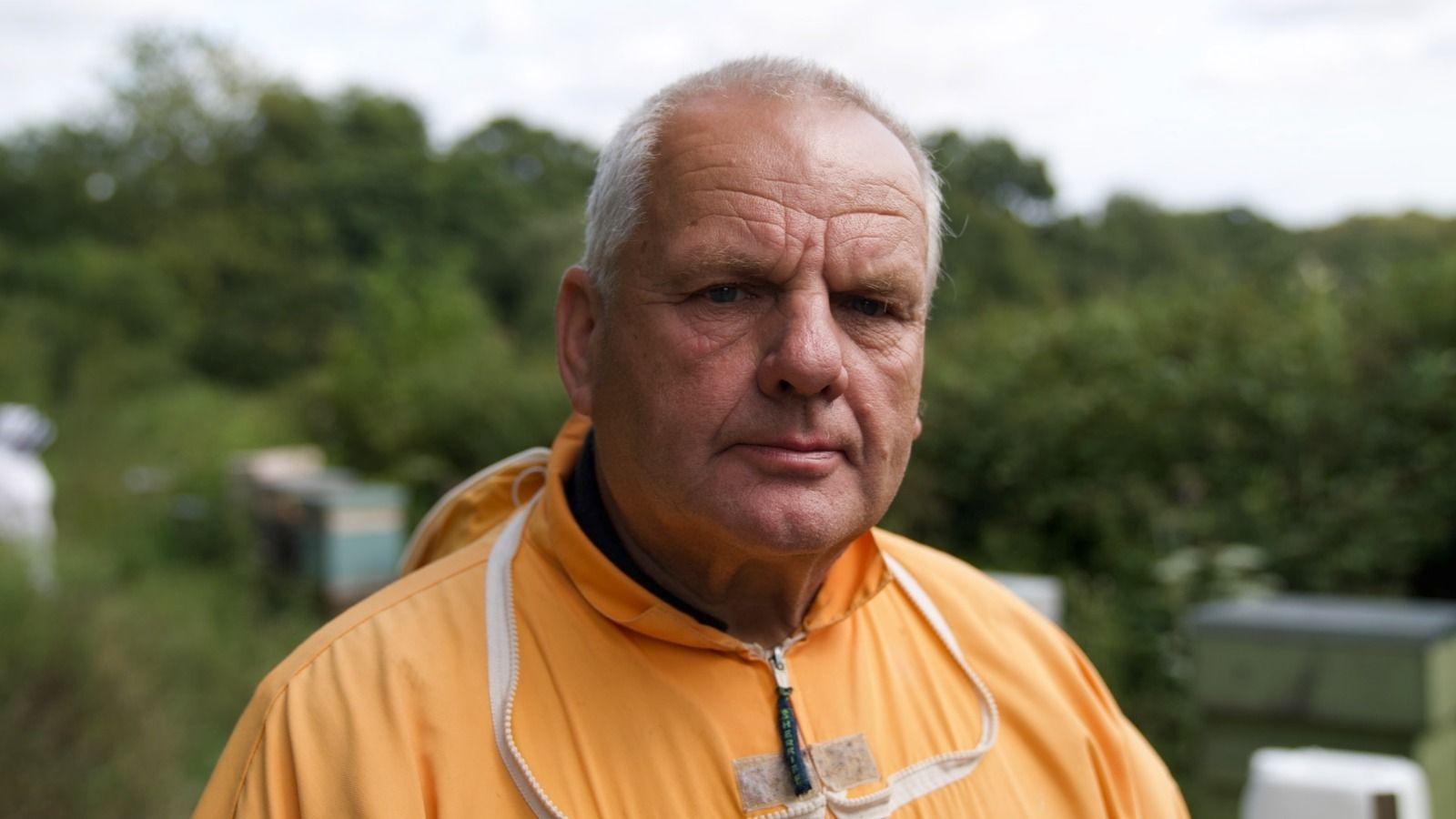 A man wearing an orange beekeeper suit with hives and trees in the background.