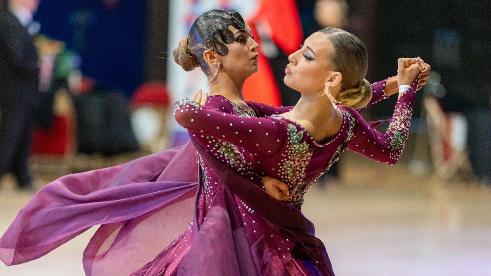 Two women holding each other in a dance position. They are both wearing purple dance dresses embossed with sequins