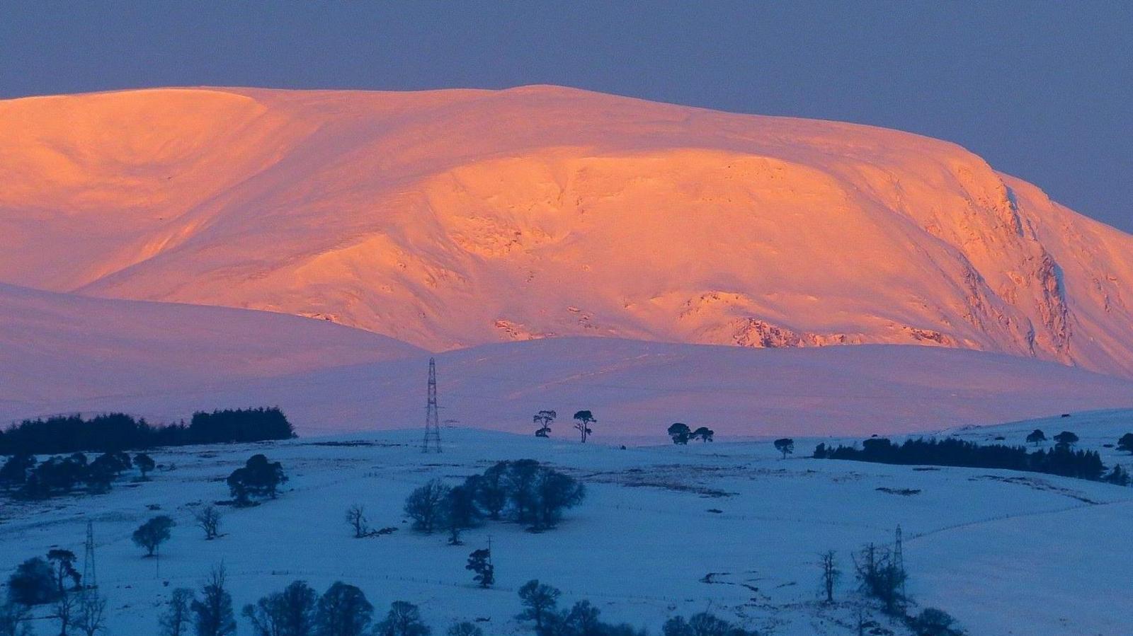 Snow-covered Ben Wyvis mountain glows pink and orange in the light of the sunrise. Fields in the foreground are in shadow and a chilly blue colour.