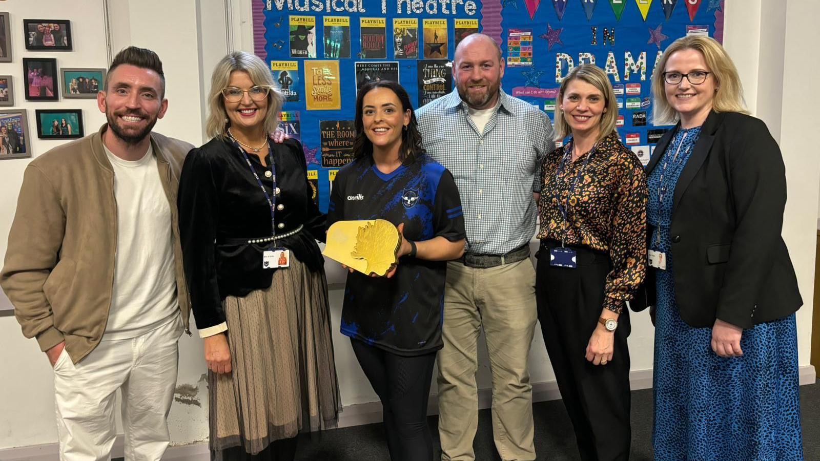 Ciara Mulholland, who has dark hair, and is wearing a school tracksuit, stands along with teaching colleagues  and One Show presenter Ryan Hand. She is holding an award.