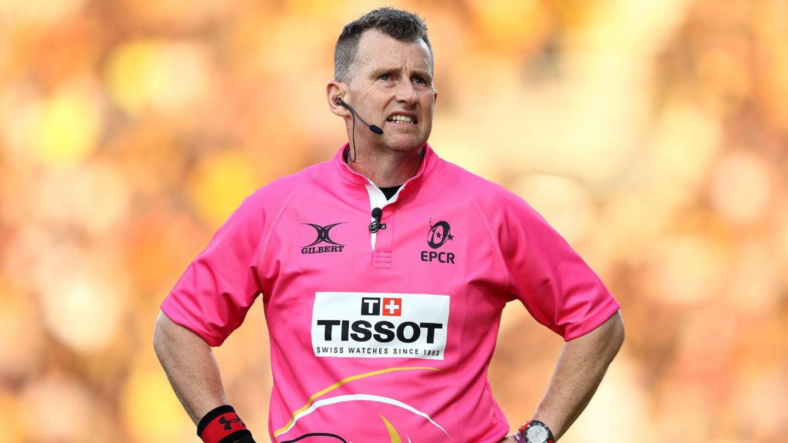 Nigel Owens in a pink rugby match jersey. He has his hand on his hips and is talking into  his earpiece microphone 