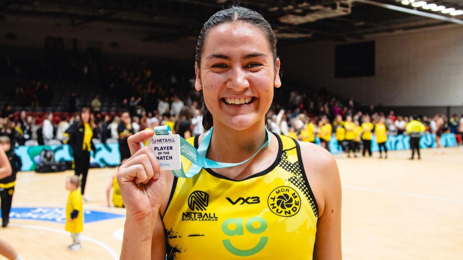 Sophie Fawns, smiling in Manchester Thunder's yellow netball outfit, holds her Player of the Match badge, with a netball court in the background