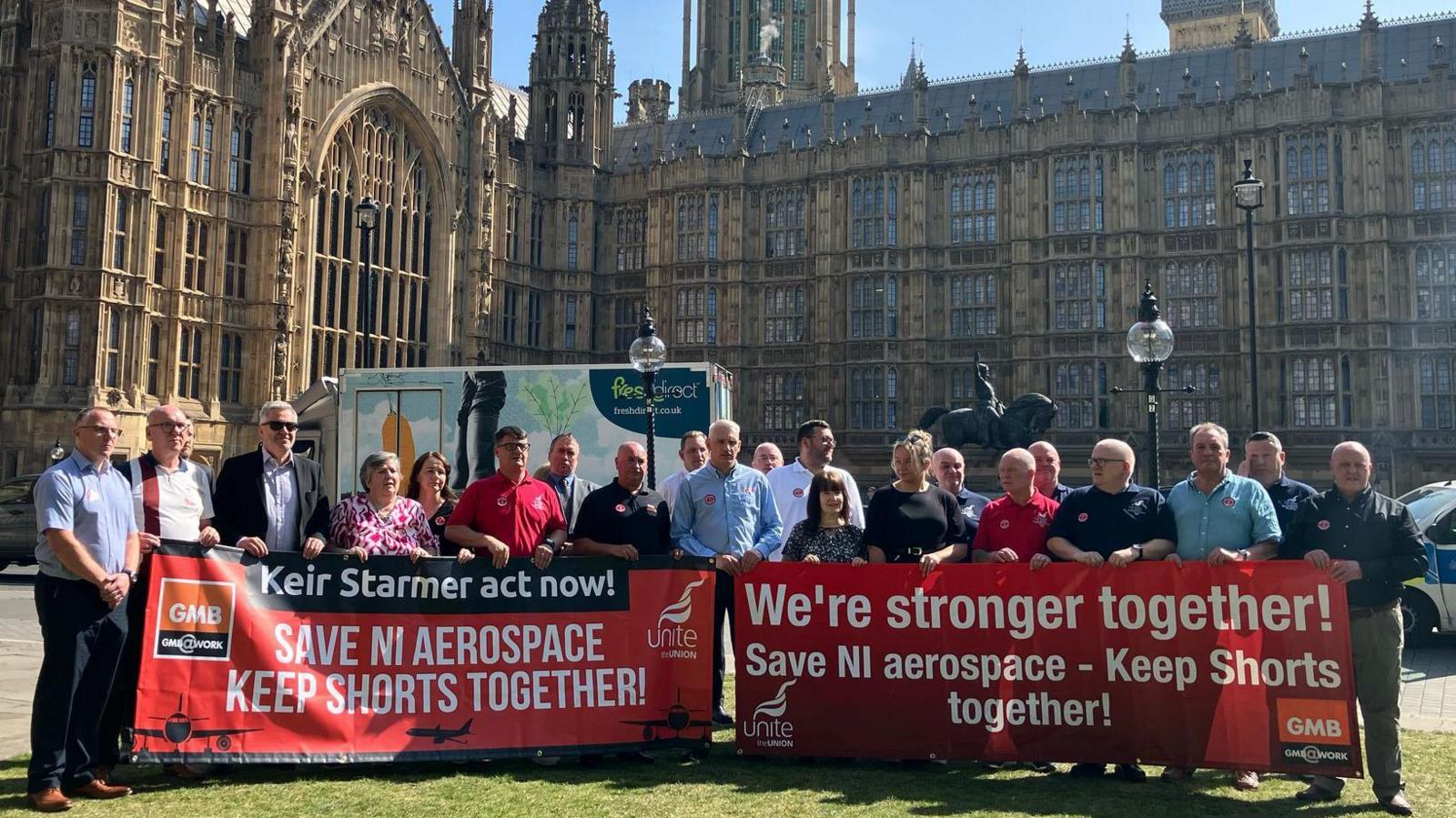 A group of protesters, stood outside Westminster, holding red GMB union banners that say "Keir Starmer act now! Save NI Aerospace keep shorts together... We're stronger together, save NI aerospace"