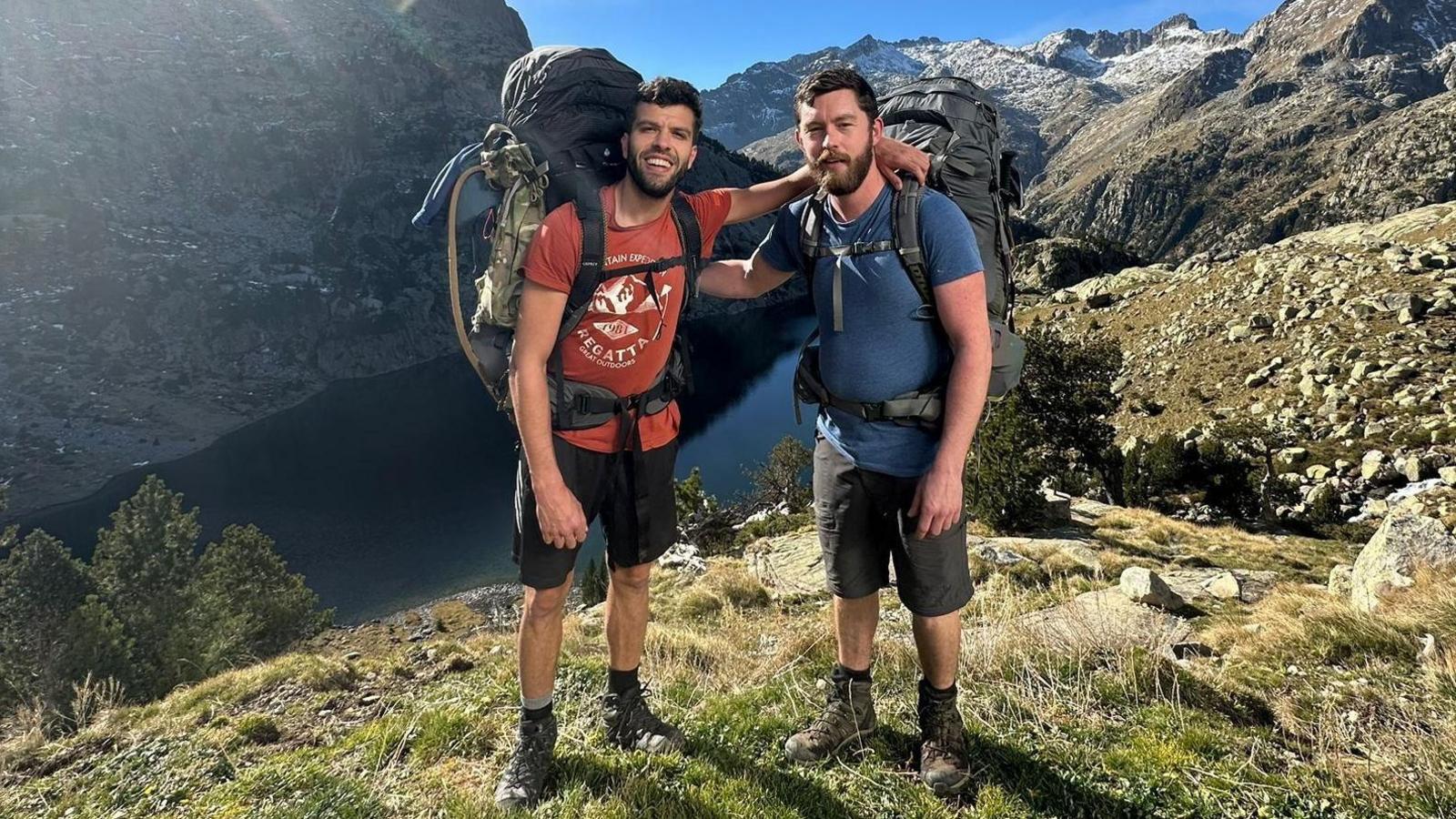 Two young men in shorts, T-shirts with large backpacks in front of a lake surrounded by mountains.