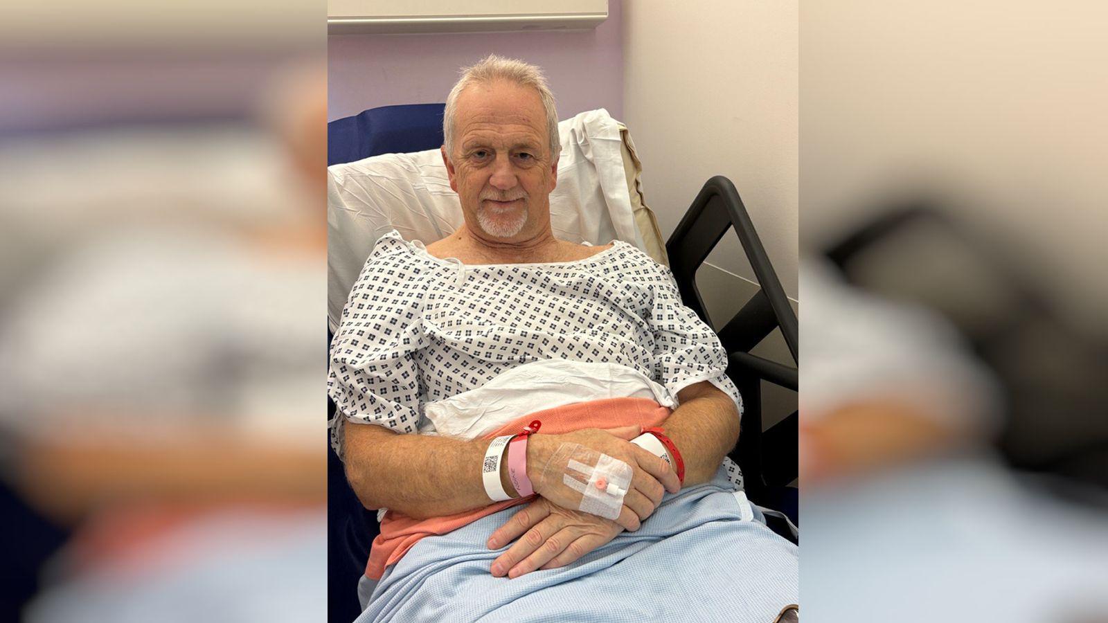 A man in a hospital bed. He is awake and smiling.