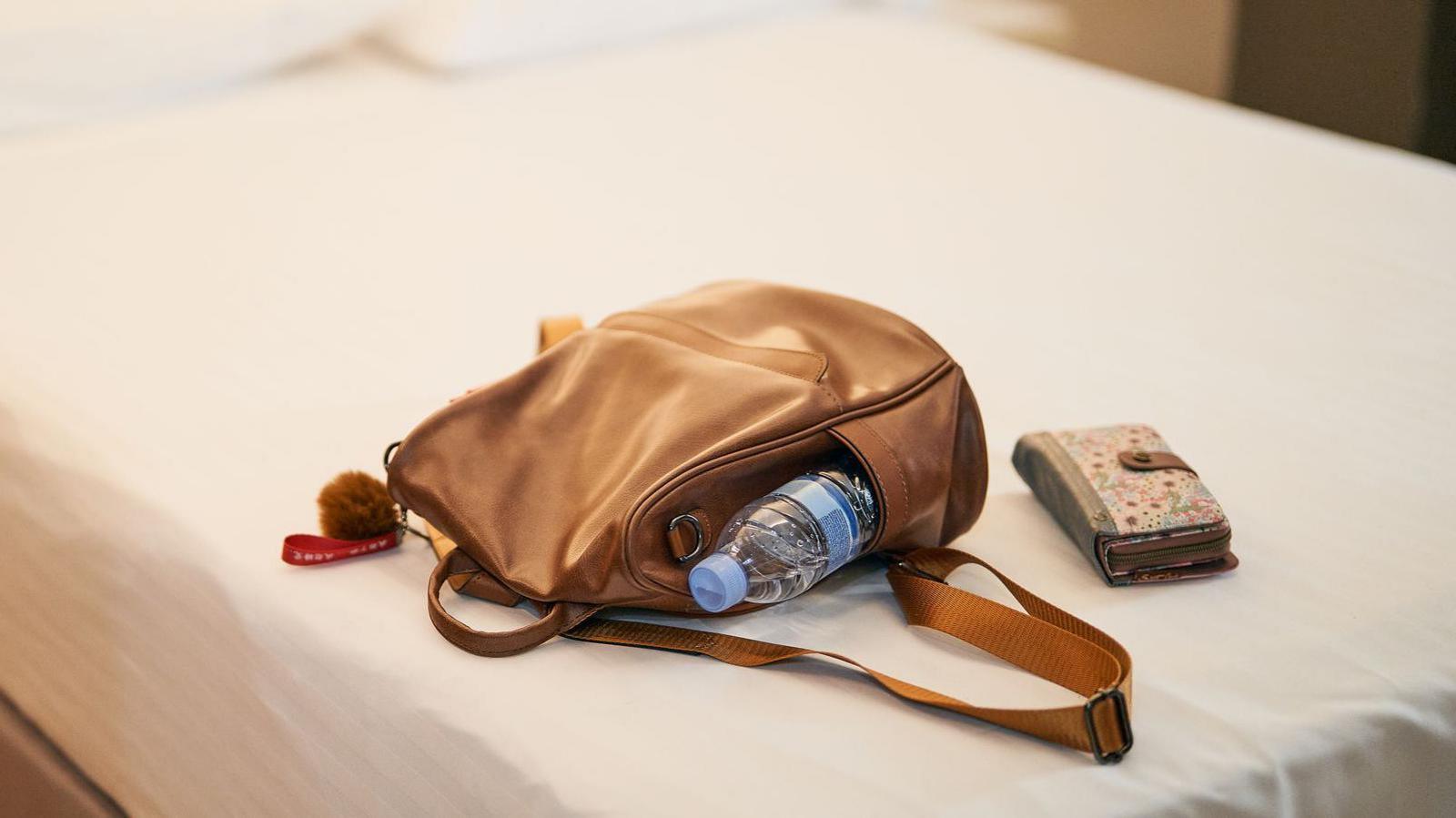 Brown leather backpack with a water bottle and wallet resting on a made bed in a hotel room.