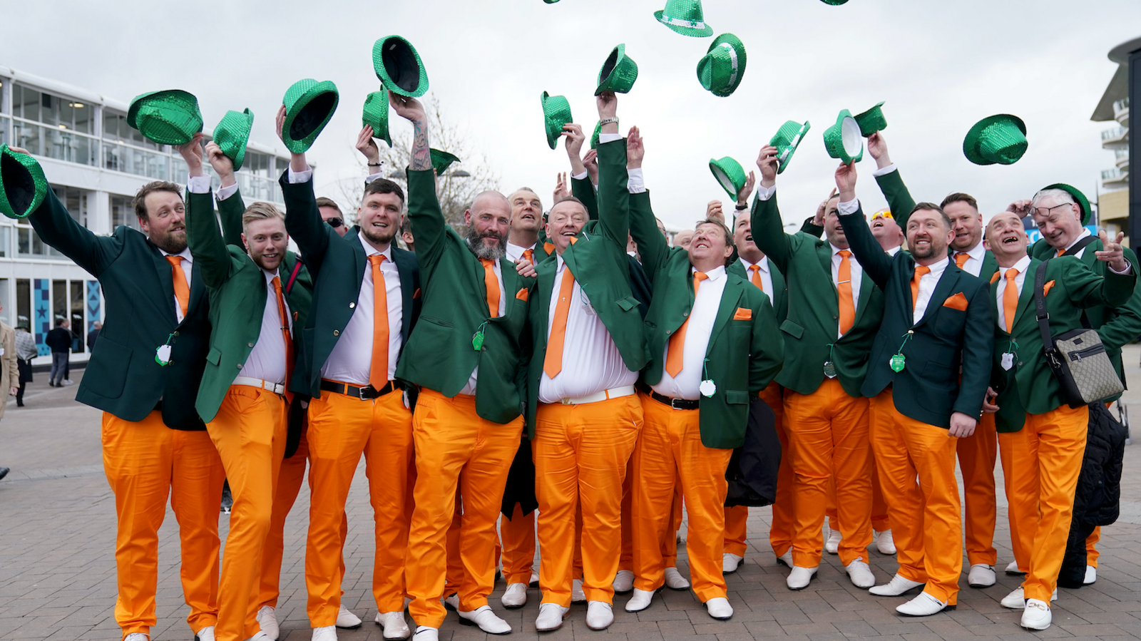 Men dressed in orange trousers, with green jackets. They have orange ties and pocket squares. All of them are smiling and throwing their green hats in the air.