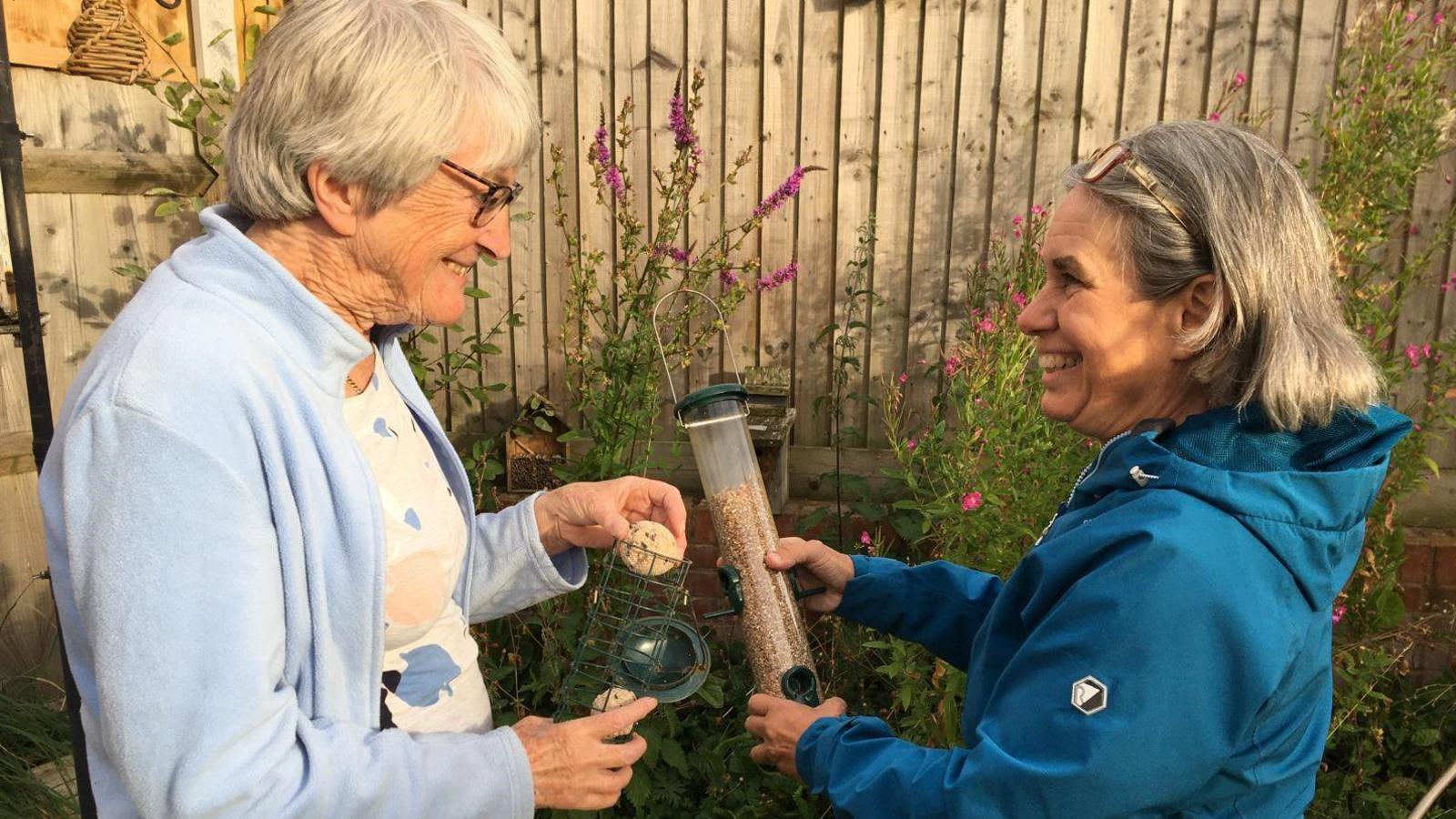 A woman with short grey hair and black glasses is wearing a light blue jacket and white patterned top. She is putting a fat ball into a bird feeder. Next to her is a woman with a grey bob. She is wearing a blue jacket and holding a bird feeder with seed in it.