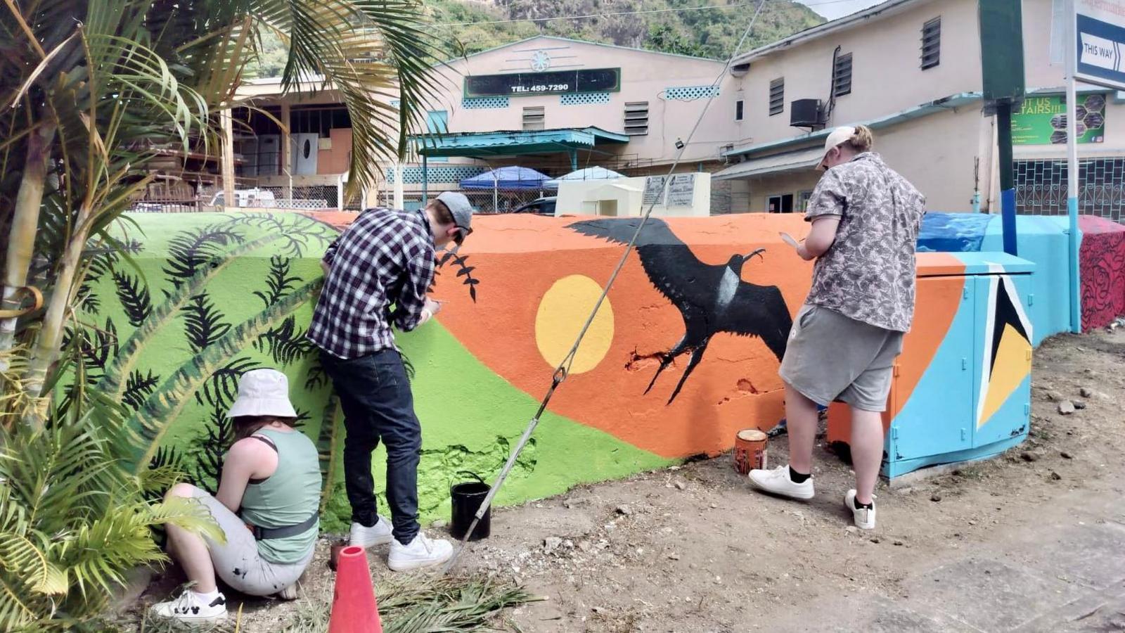 A young woman and two young men, wearing casual clothes and with the backs to the camera, paint a mural on a wall in a community in Saint Lucia. The mural uses bright shades of green, orange and blue and features a black bird flying by a yellow sun. Palm trees stand to the left.