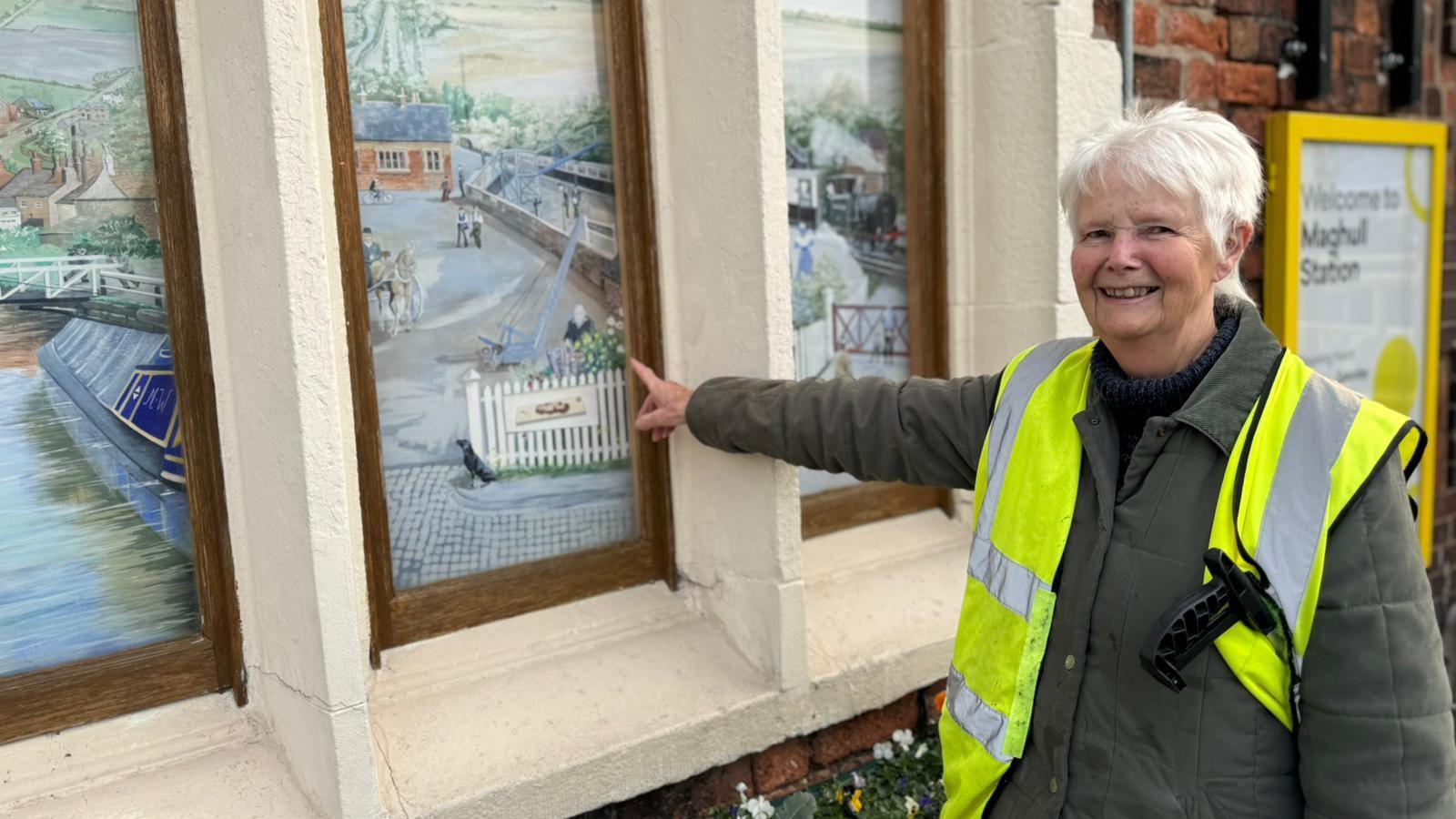 Margaret Walton, who has short white hair and wears a dark green coat with a high vis jacket. She has a litter picker under her left arm and smiles at the camera as she points at a mural depicting the platform at Maghull. 