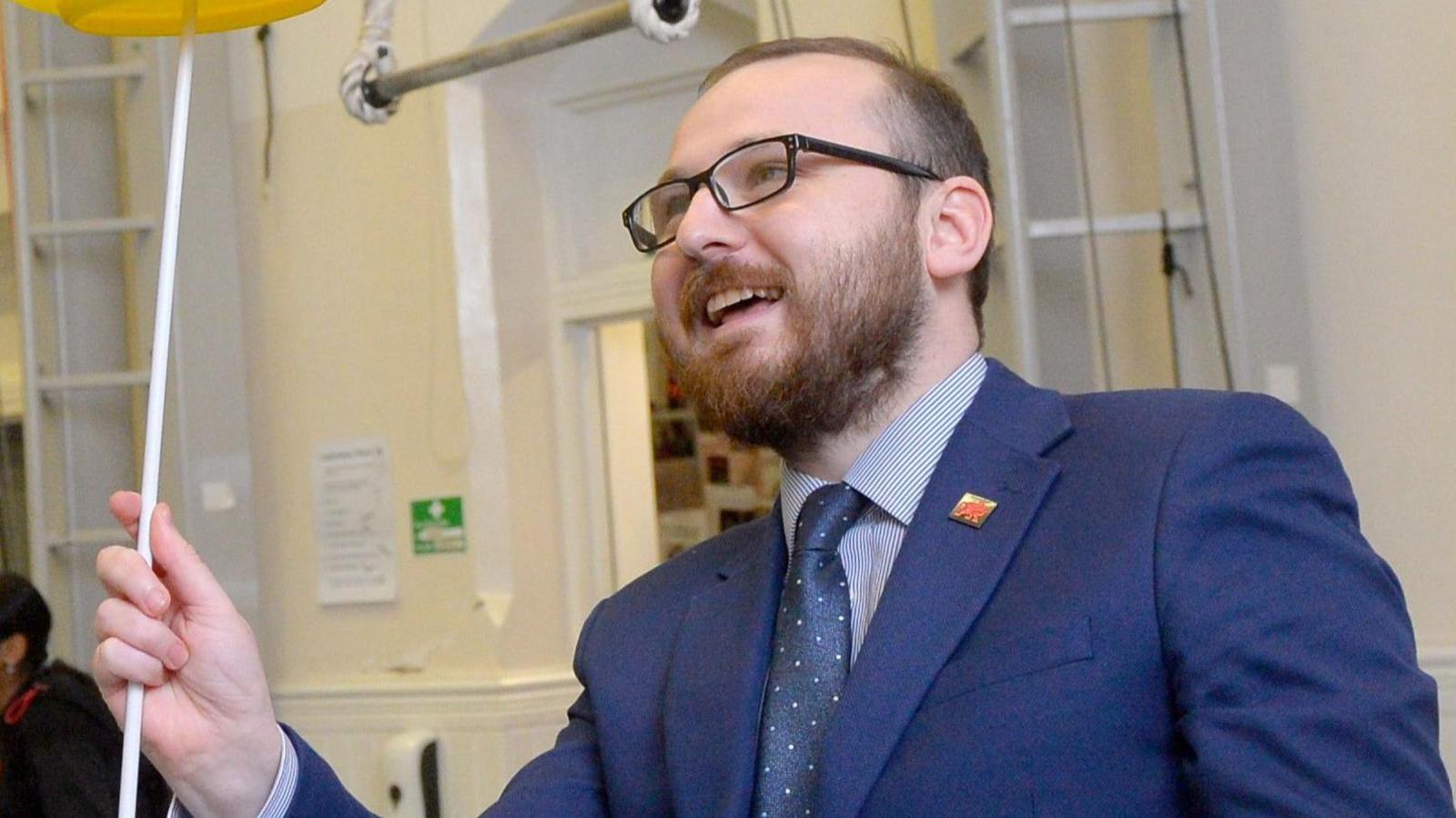 Jack Sargeant, wearing a blue suit and glasses, stood spinning plates at a circus training centre in Cardiff.