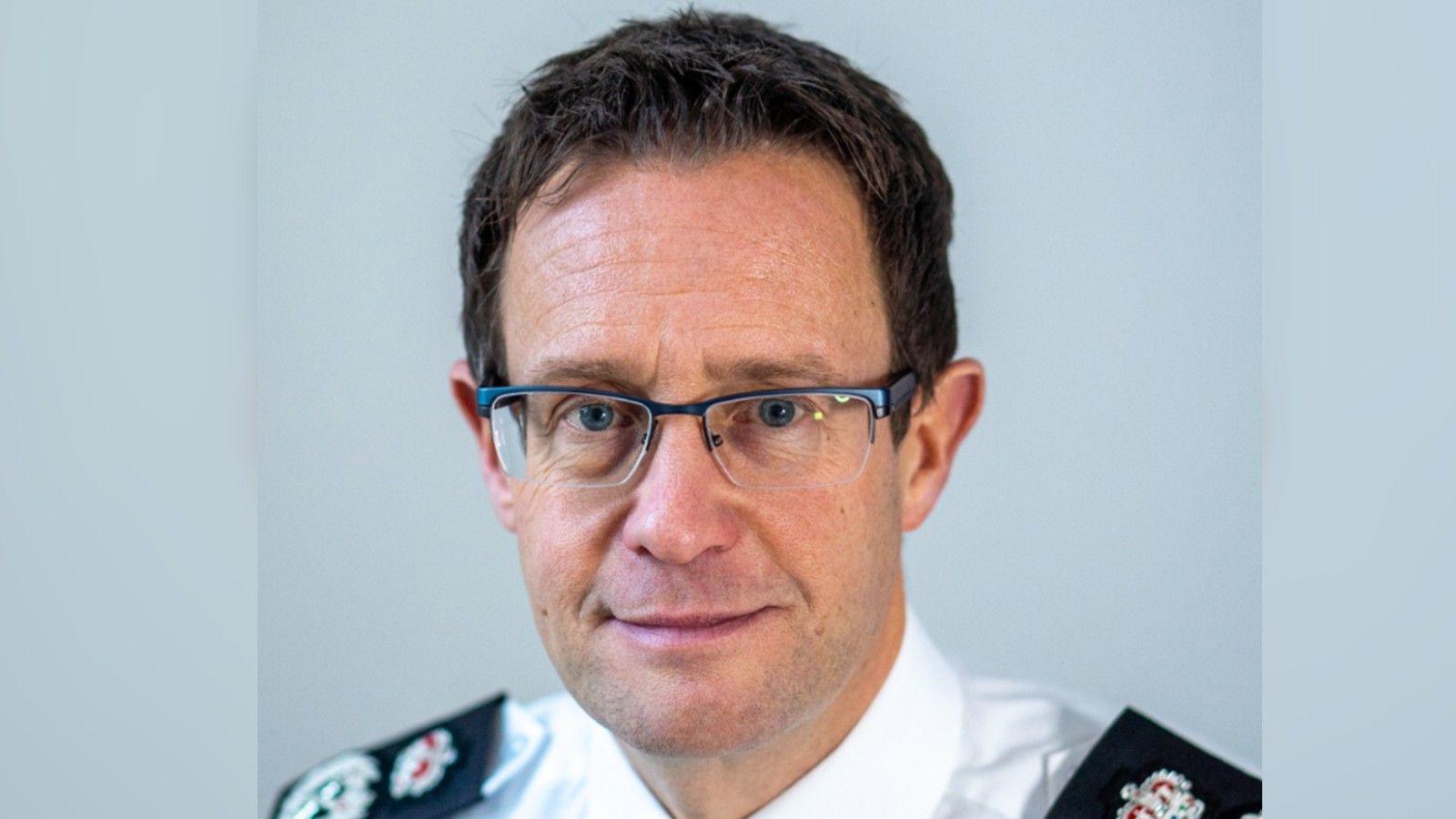 Pictured is Chris Noble against a pale blue background. He is slightly smiling, wearing rectangle frame glassed and has brown hair and blue eyes. He is wearing a white shirt with black shoulder trim and police badges.