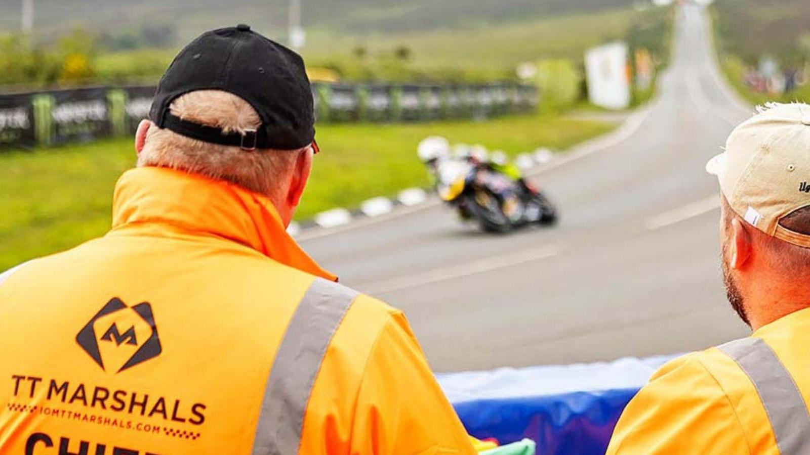 Two men in orange coats and baseball caps look on at the race course from behind a blue crash barrier as a motorcycle passes by quickly on a sharp bend.