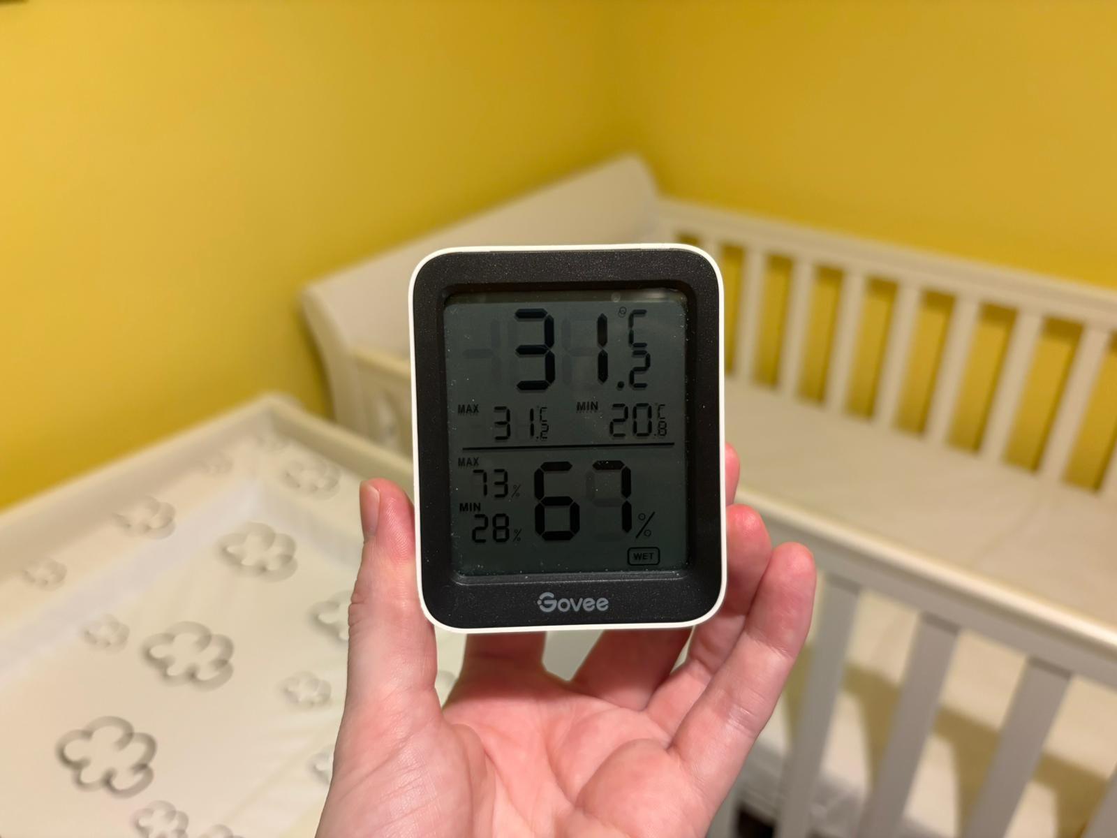 A digital thermometer is being held up in front of a crib. It shows a temperature of 31.2C