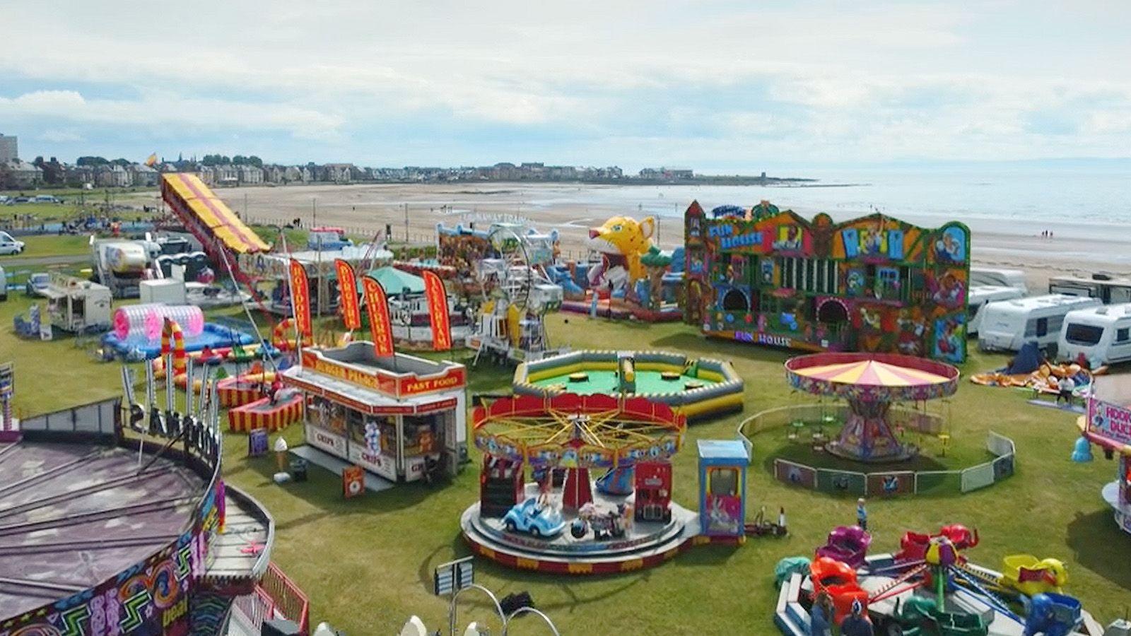 A seaside view of funfair rides, with a beach and coast in the distance under blue sky.