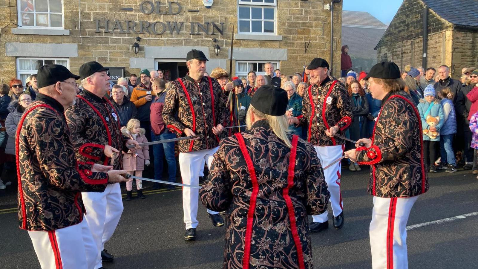 A group of sword dancers wearing black caps and traditional jackets - black, with an elaborate paisley design in shades of red and white - dancing with their swords outside a stone-built pub.