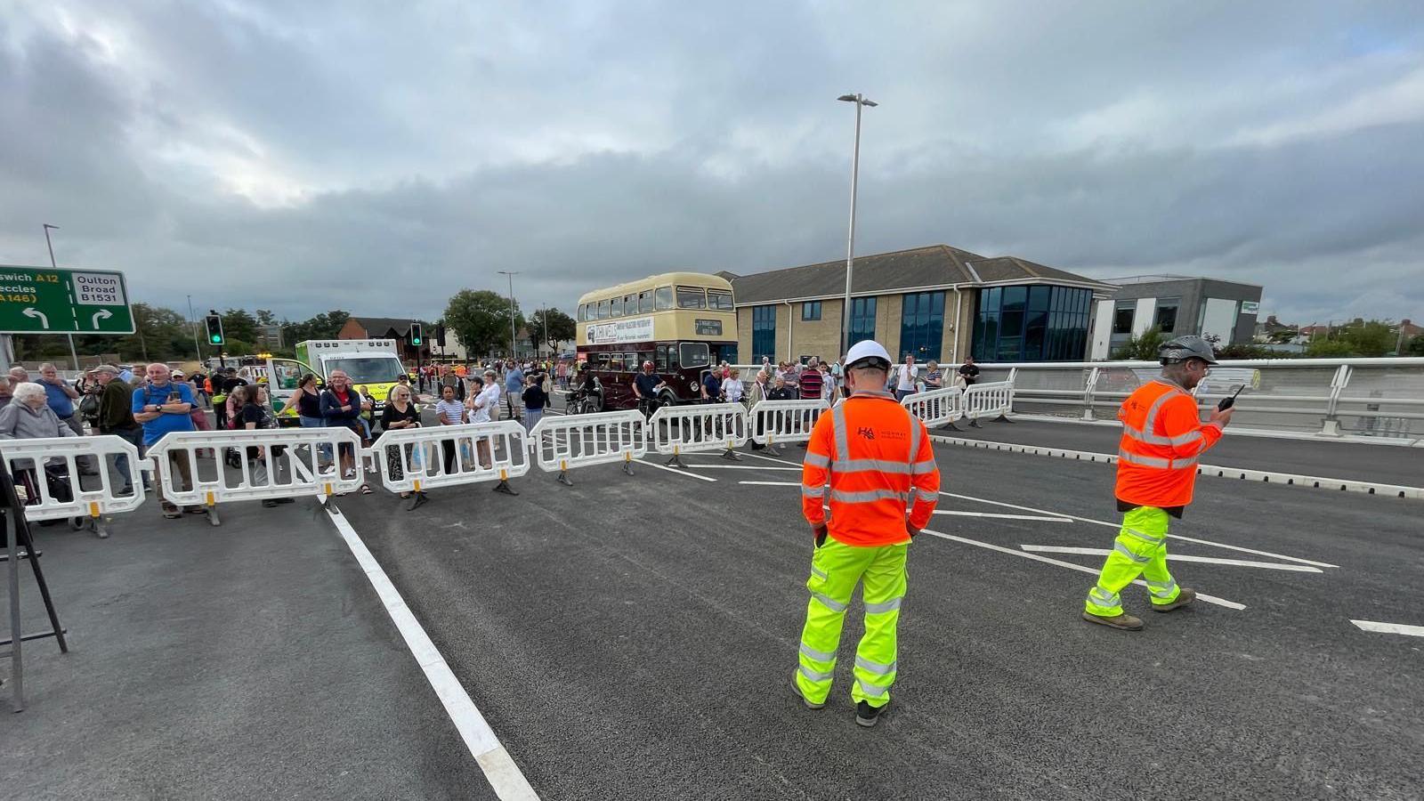 Two highways workers in high viz clothing stood in front of a barrier with people waiting behind