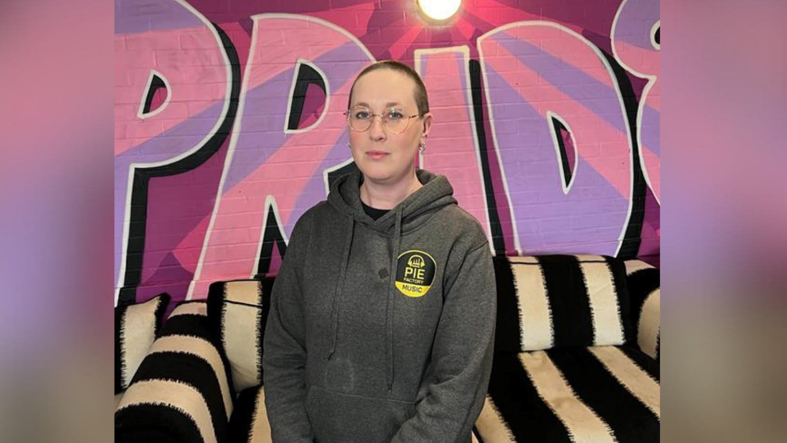 CEO of Pie Factory Music, Zoe Carassik, wearing a grey hoodie and stood against the backdrop of the Pie Music Factory charity headquarters in Ramsgate. 'Pride' is painted in pink and purple on the wall and Zoe is stood in front of a black and white striped sofa.