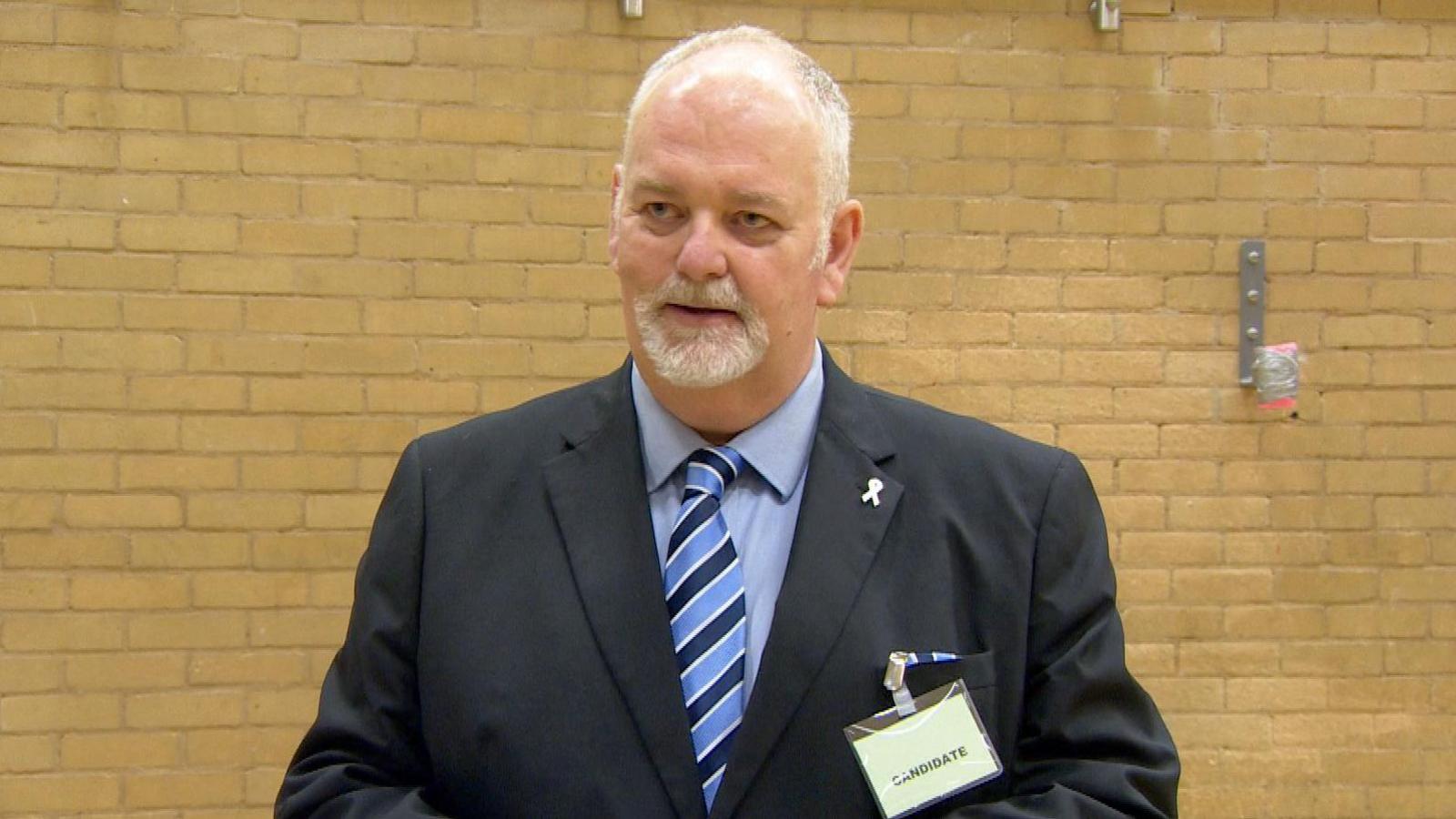 A man with white hair and beard at the recent election count. He is wearing a suit.