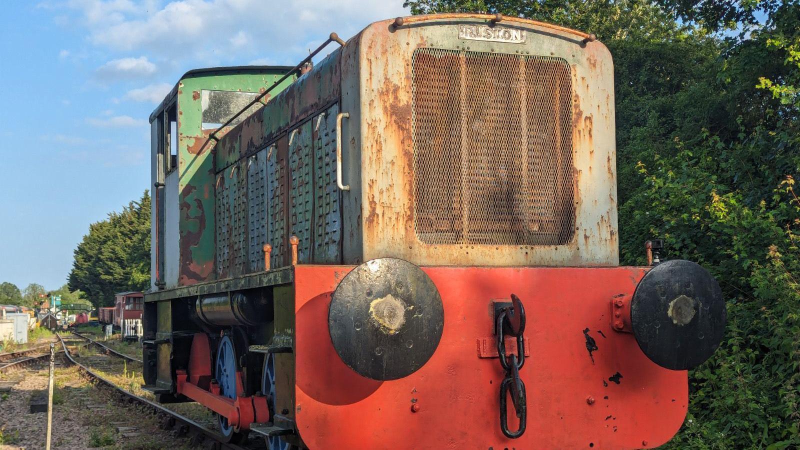 A close up view of the front of a large locotmotive. It has a lower red front with a grill above this. A cabin can be seen further back behind the engine compartment. Tracks can be seen running behind the locomotive. 