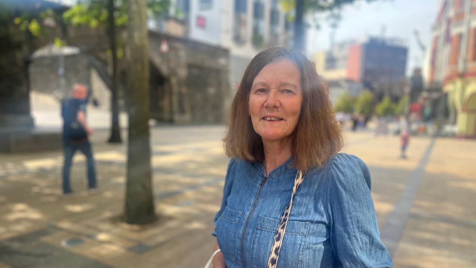 A smiling lady stands in Derry's Guildhall square. The city's walls are seen in the background as are a number of shops. She is wearing a denim top and has long brown hair. A strap of a handbag can be seen over her shoulder