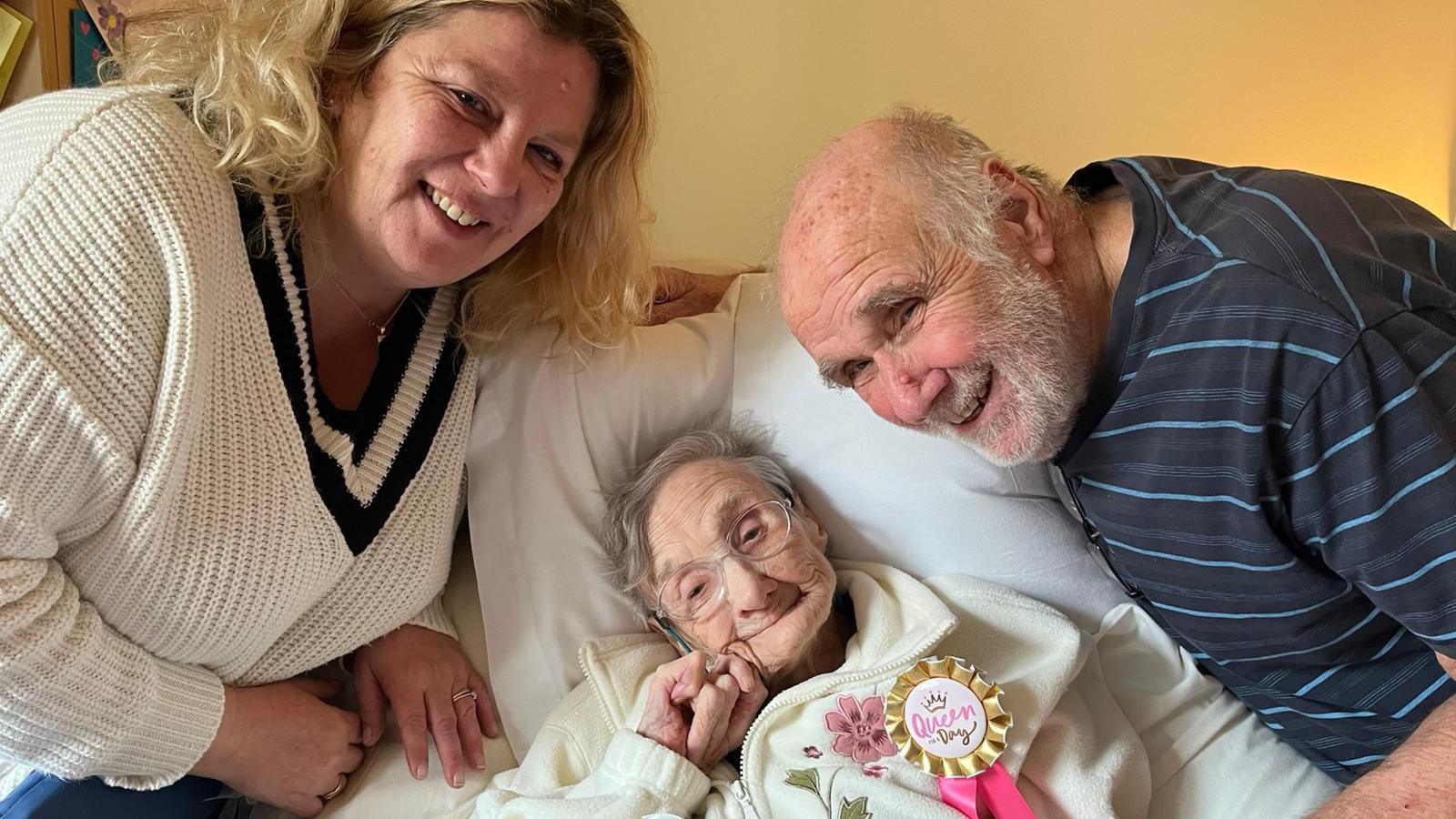 An elderly woman with short grey hair and glasses lies propped up in bed. She is wearing a dressing gown and a rosette. A younger but still relatively elderly man with short grey hair on the sides of his head and a grey beard and a younger woman with shoulder-length blonde hair lean over her, smiling at the camera.