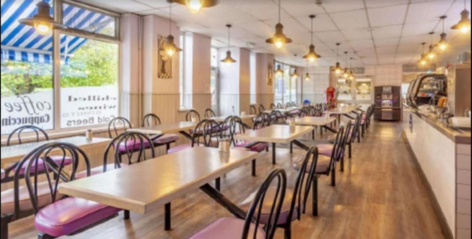 A long room is filled with dining tables and chairs. On one side of the room there are windows looking out on to trees and a blue and white striped awning can be seen. On the right side of the room there is a long bar with what appear to be food display cabinets on it.