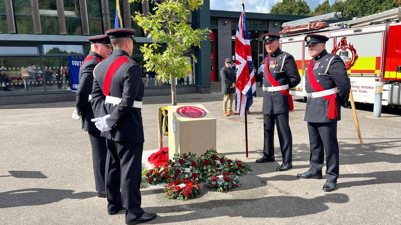 Four fire officers wearing formal dress stand around a red plaque, with wreaths placed at the foot of the plinth, and a fire engine in the background.