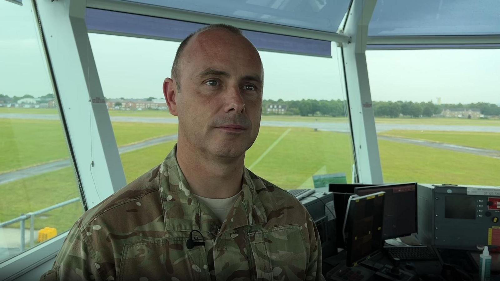 Sqn Ldr Tim Clayton speaks to a reporter who is behind the camera. He is in front of the windows of the visual control room. The green air field can be seen though the windows. 