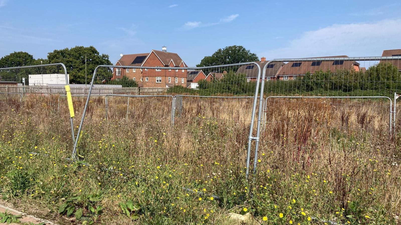 A metal fence in an overgrown area of grassland while in the background homes can be seen.