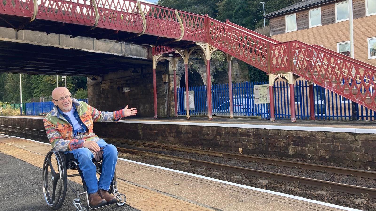 A man in a wheelchair on a platform gesturing to the other side which appears to be only accessible via steps up to a footbridge 